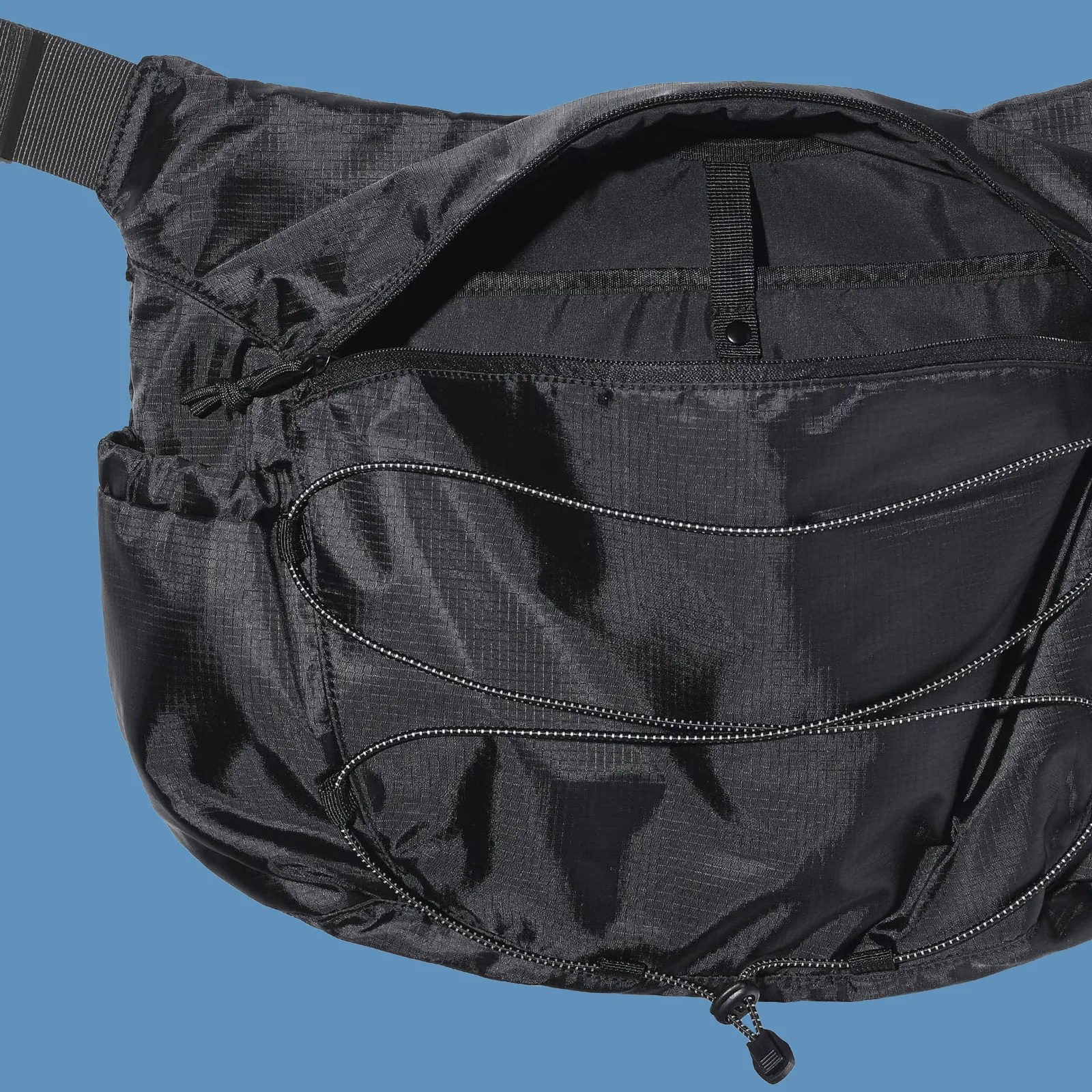 Gu Ripstop messenger bag in black shown empty and laid flat with main compartment open to reveal its interior pockets against a solid blue background