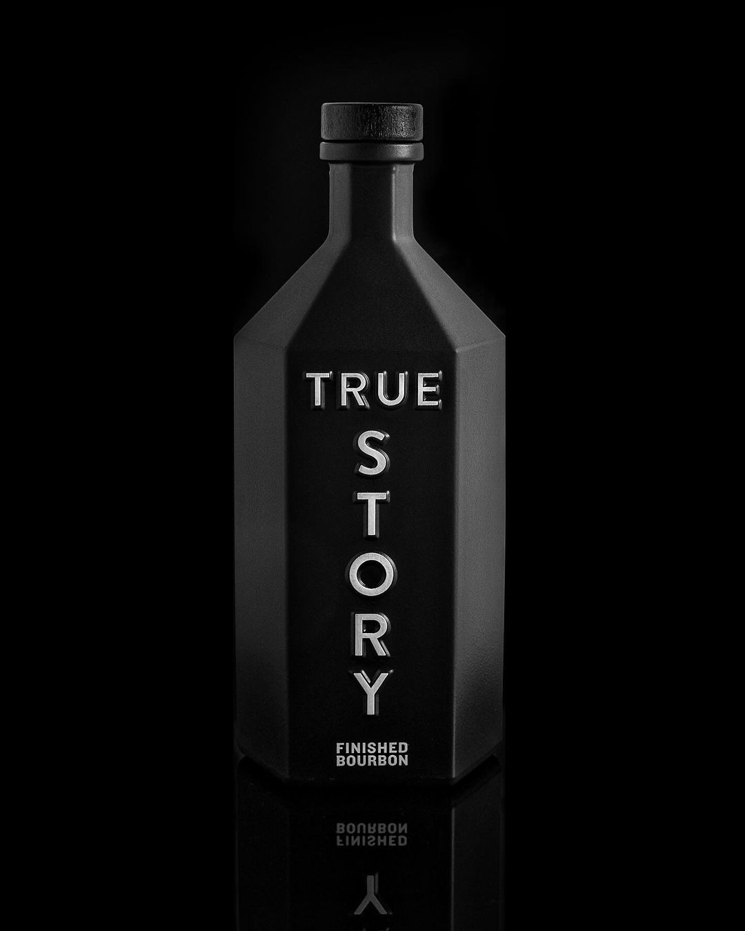Two bottle of True Story Bourbon shown head on. The bottle is all black and hexagonal shaped with black bold type. 