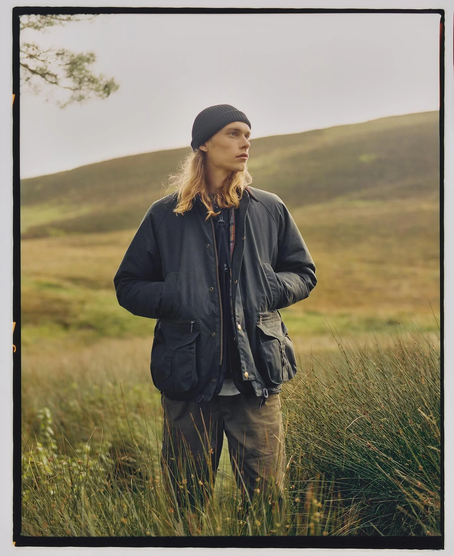A white male with long blonde hair wearing a beanie and Barbour’s Modified Beaufort waxed canvas jacket while staring into the distance standing in a field of long green grass in front of a sloping green hill. 