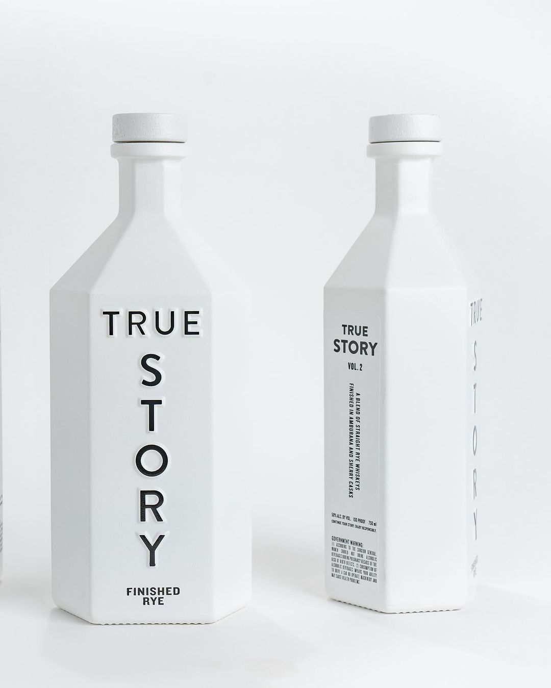 Two bottle of True Story Finished Rye shown from different angles. The bottles are all white and hexagonal shaped with black bold type. 