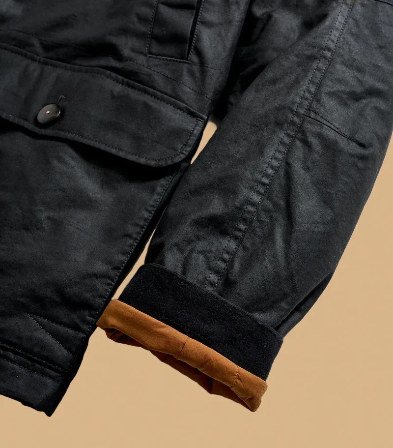 A close up of the sleeve cuff and bottom right corner of the Billy Reid Hunting Jacket laid flat and zipped up on a light tan background. The image highlights the lighter brown interior lining of the sleeve cuff that's shown rolled up. 