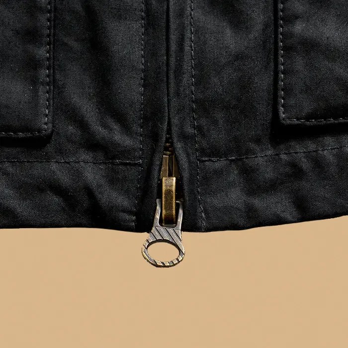 A close up of the distressed antique brass main zipper on the Billy Reid Hunting Jacket. The close up highlights the diagonal stripped textured detailing on the zipper. The image background is a light tan color. 