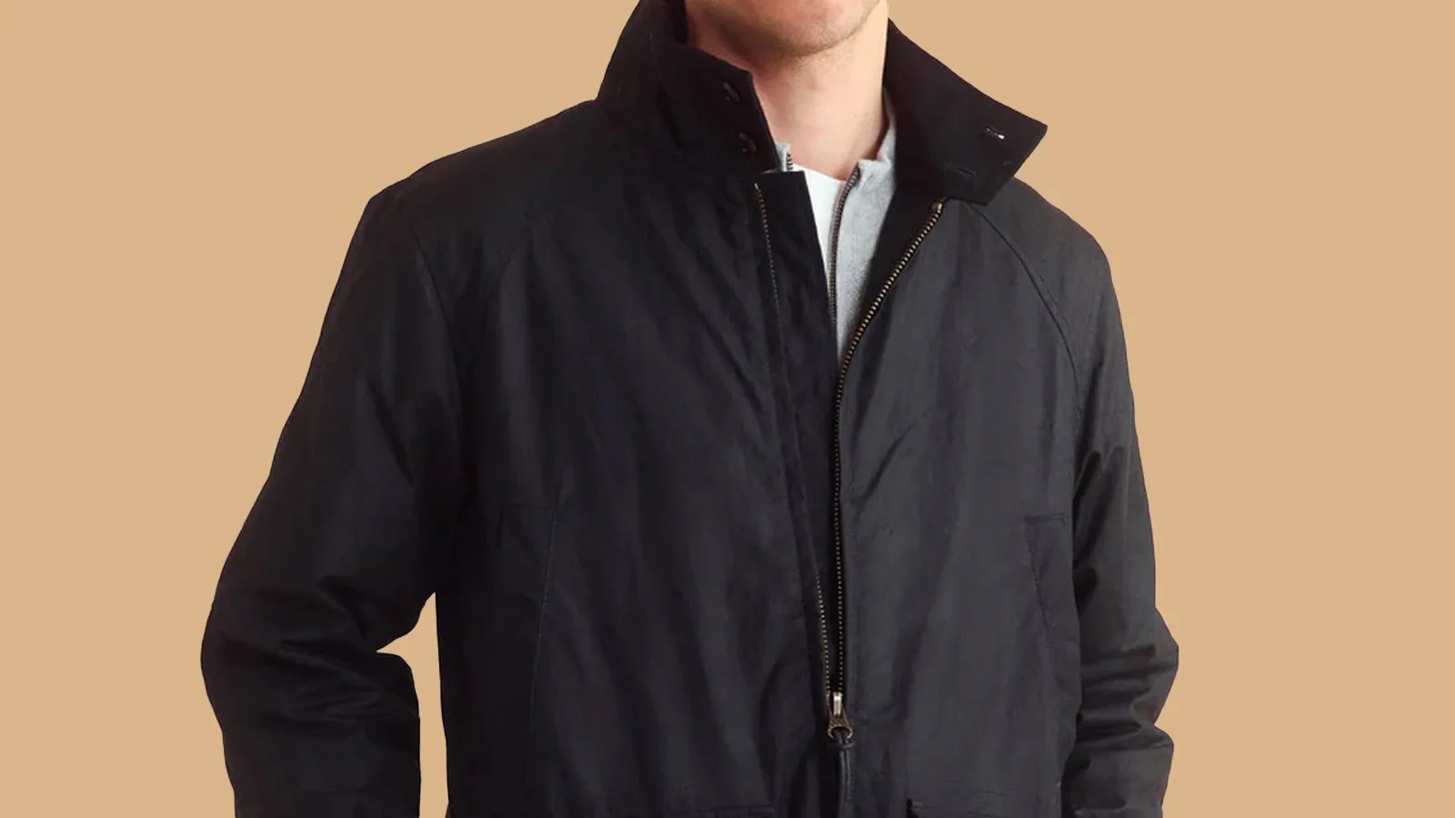 A cropped image of a male model wearing a Billy Reid waxed canvas hunting jacket. The blacked waxed canvas jacket is shown from right above the models waist to the models neck. The central zipper is slightly unzipped at the top and the collar of the jacket is popped up.