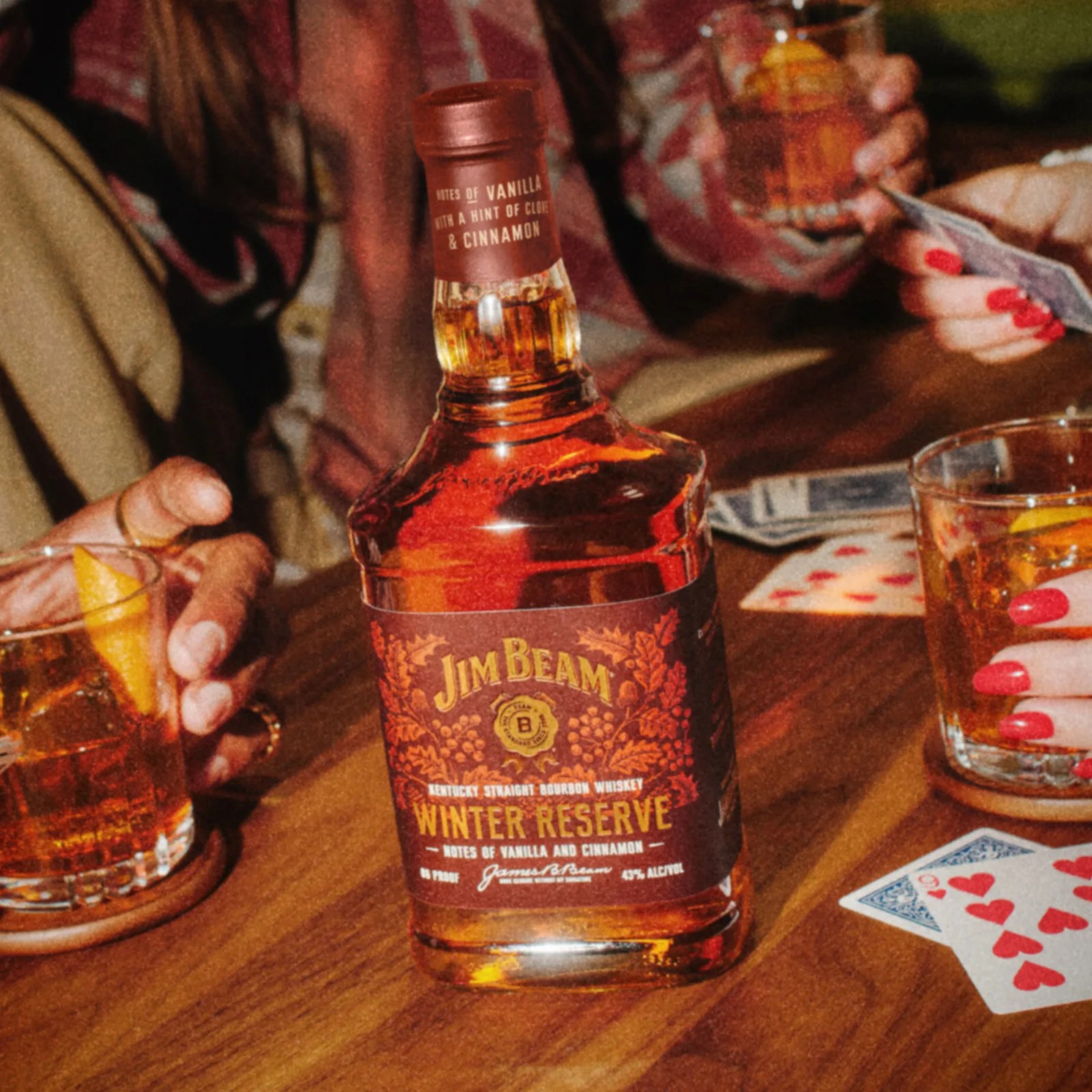 A bottle of Jim Beam Winter Reserve sitting on table surrounded by playing cards and various drinkers holding full glasses
