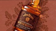 A close up of a bottle of Jim Beam Winter Reserve Bourbon shown at an angle highlighting the dark maroon brownish label. It's sitting on a dark read background with a holly leaf pattern.
