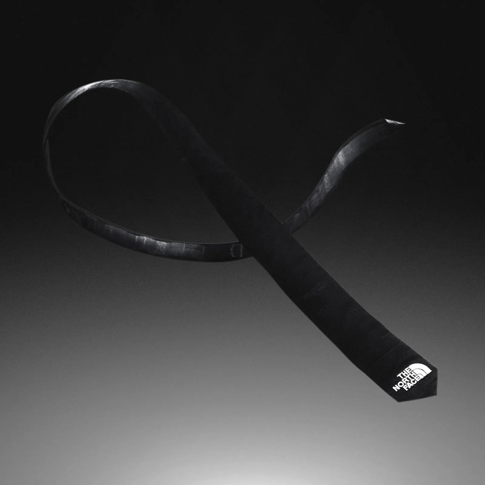 The North Face Remade Tech Tie, which is a black skinnny tie with The North Face logo at the tip shown floating and twisted around against a black gray gradient background