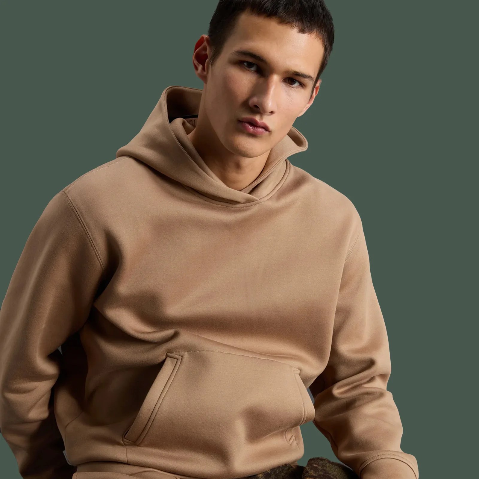 Todd Snyder x Woolrich Luxe Tech Hoodie in camel being worn on male model slouching against a forest green background