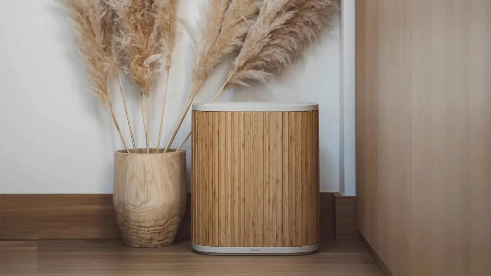 Windmill air purifier in white with a bamboo wood colored exterior sitting on a wood floor in a corner next to a light birch-colored wall and a tan vase filled with tan colored plant steams
