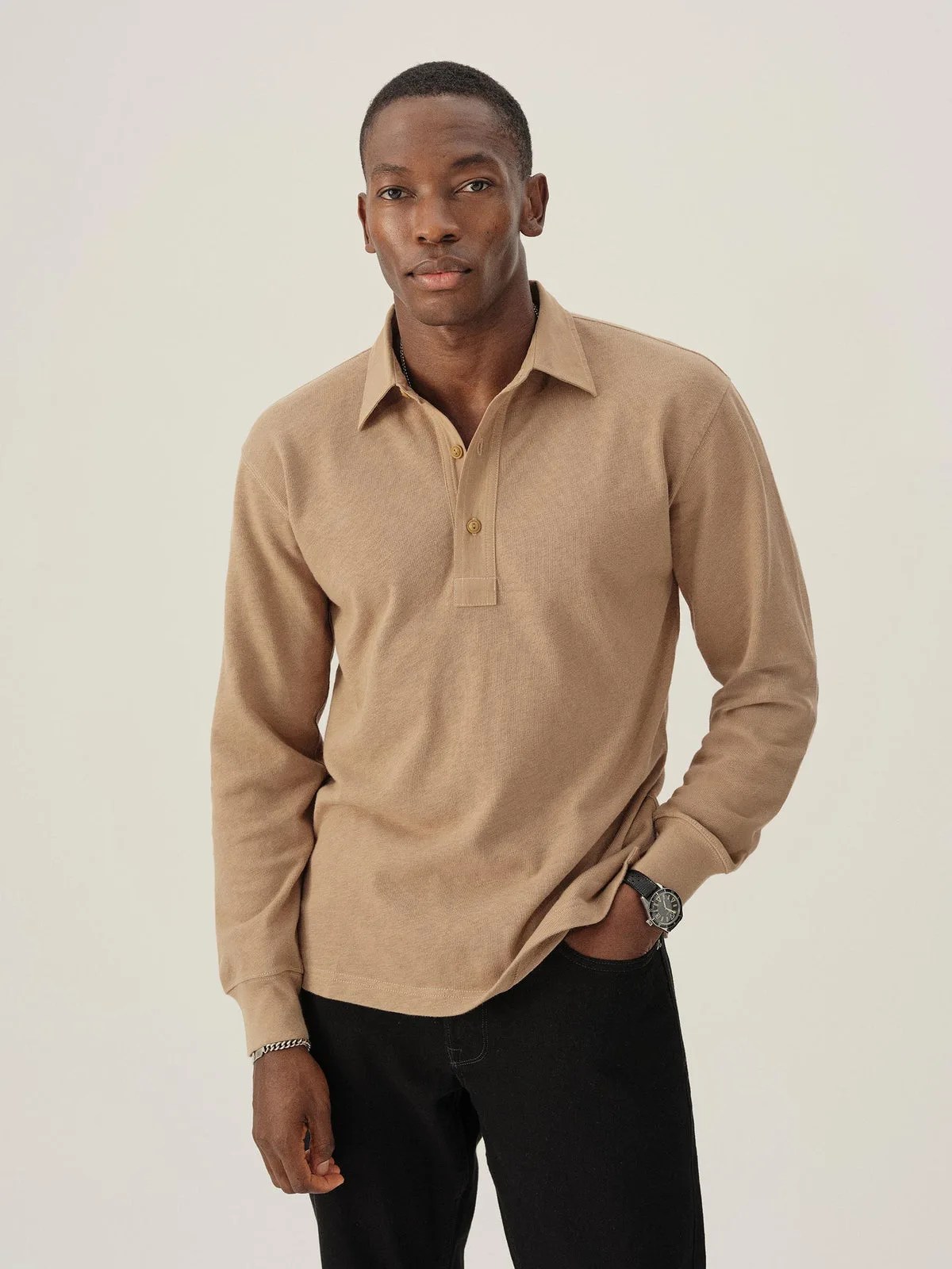 A black male model wearing Buck Mason’s Field-Spec Rugby Polo in a Sandstone color that looks like a light tan. The model is facing forward with his hand in his pocket. 