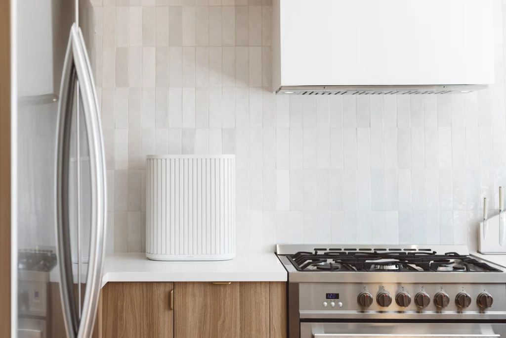 The Windmill Air Purifer in White sitting on a white countertop in a modern kitchen next to a stainless steel stove. The wall is covered in greyish white tiles. The lower cabinet is made of light wood. A set of stainless steel fridge doors can be see on the left of the photo. 
