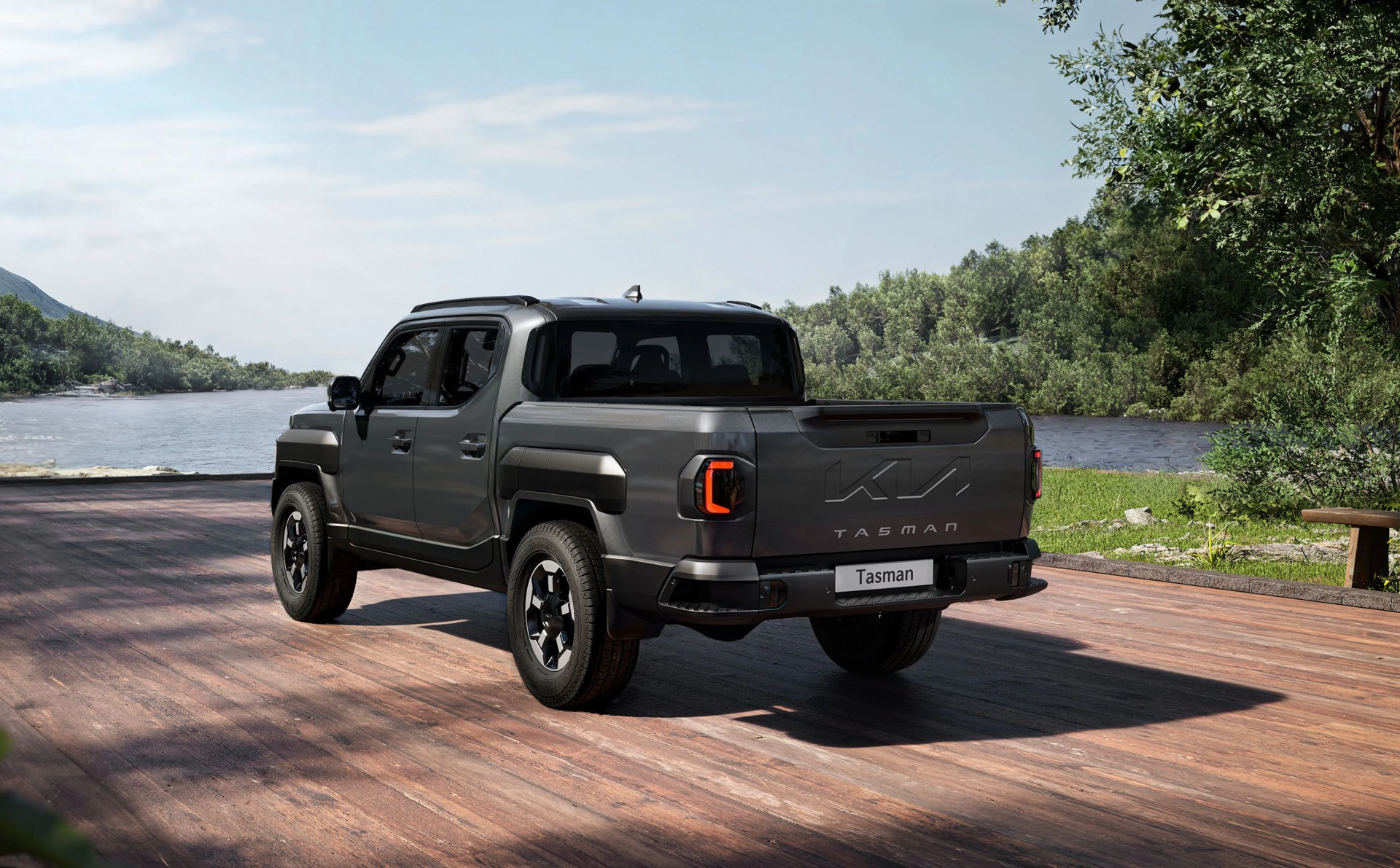 The Kia Tasman pickup truck in a dark grey color shown at an angle. The rear left corner of the truck appears closest in the image and highlights the truck’s tailgate details. The truck appears to be sitting on a wooden platform or dock next to a lake surrounded by lush green foliage. 