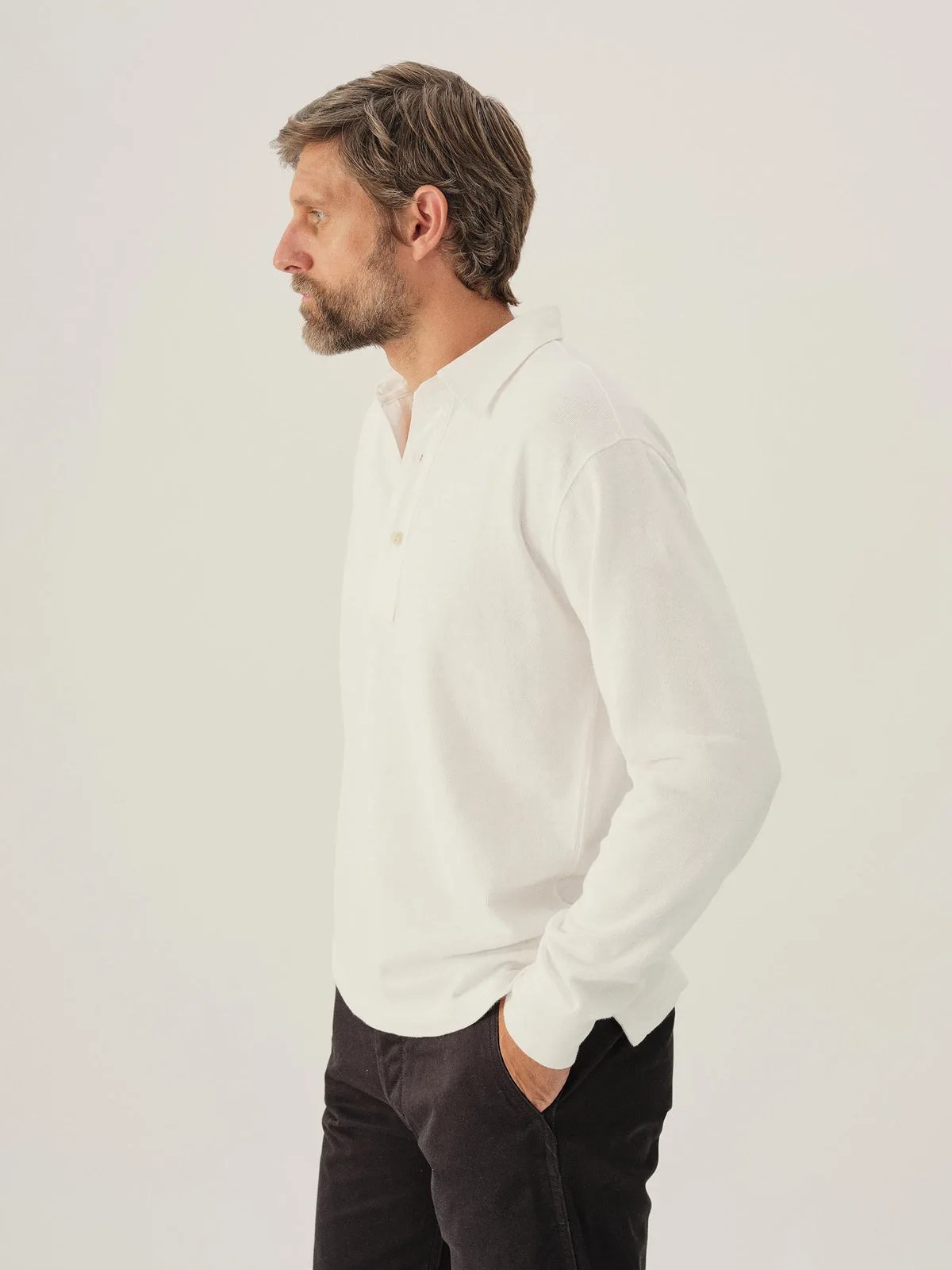 A white male model wearing Buck Mason’s Field-Spec Rugby Polo in a white color. The model is facing to the left with his hand in his pocket. 