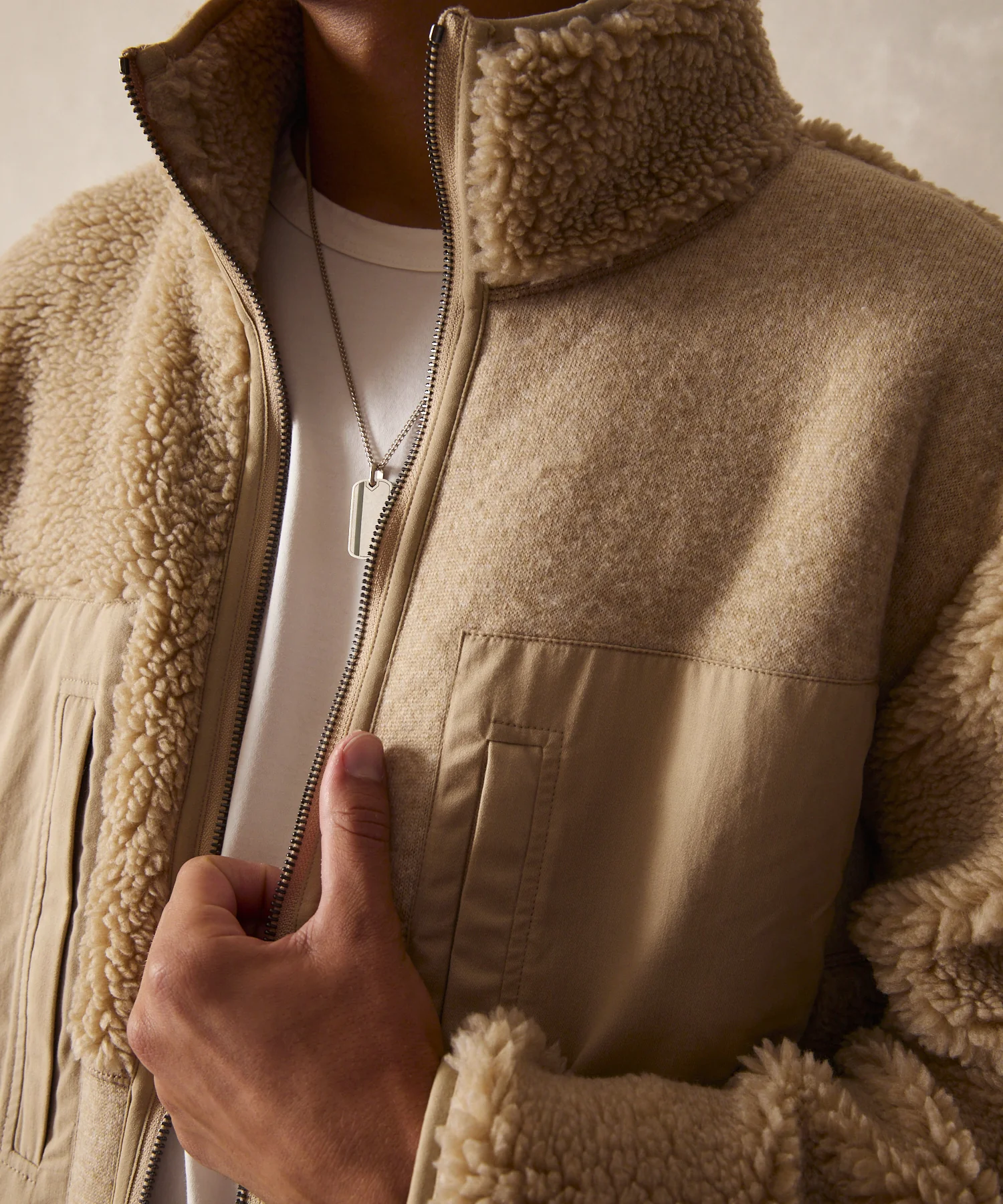 A close up image of an unzipped Todd Snyder fleece jacket in a soft Khaki color being worn by a model who is holding their hand against the open zipper line. 