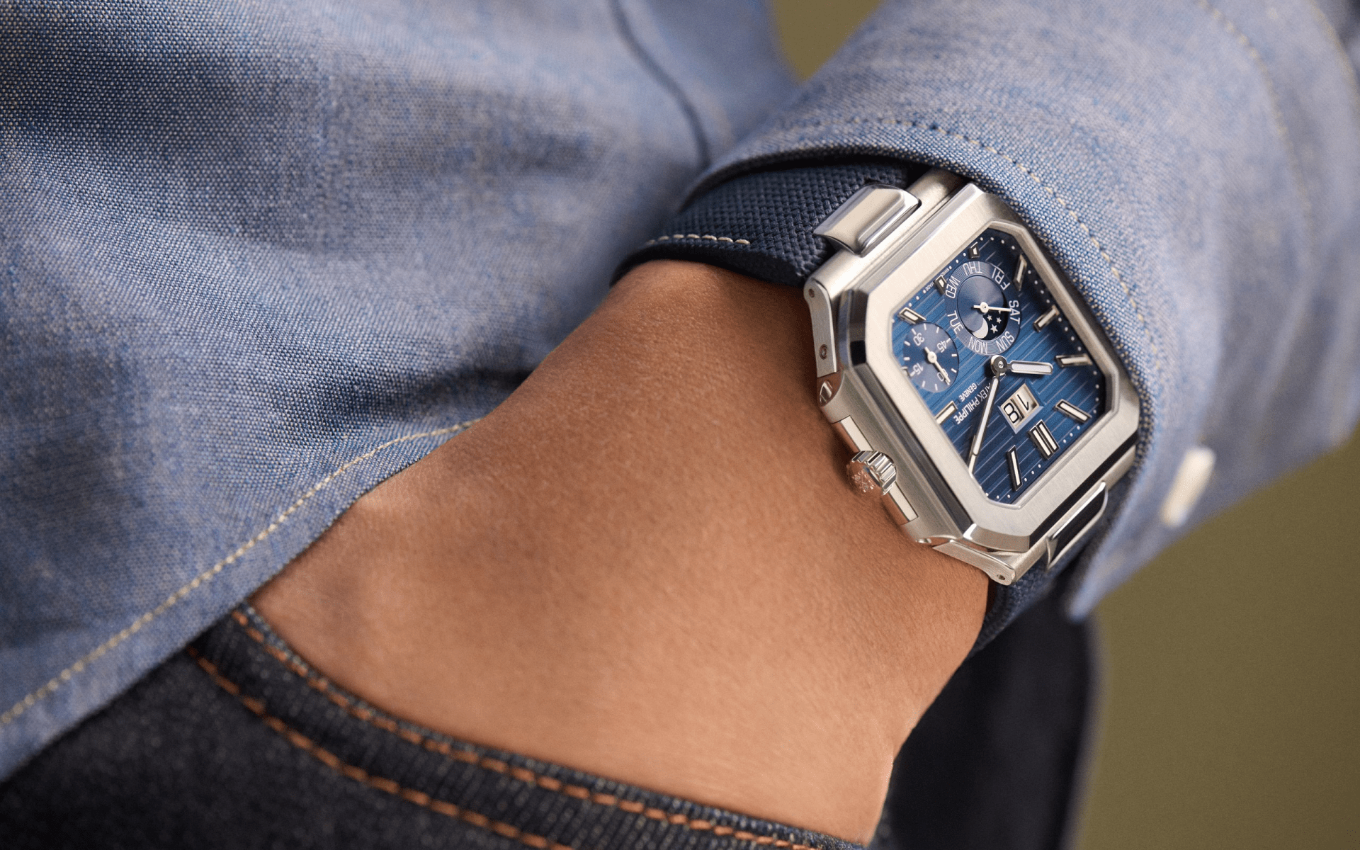 A close up of the Patek Philippe 5822_ Cubitus watch being worn on a male wearing a light blue sweater. The hand with the watch is being tucked into a jean pocket. 