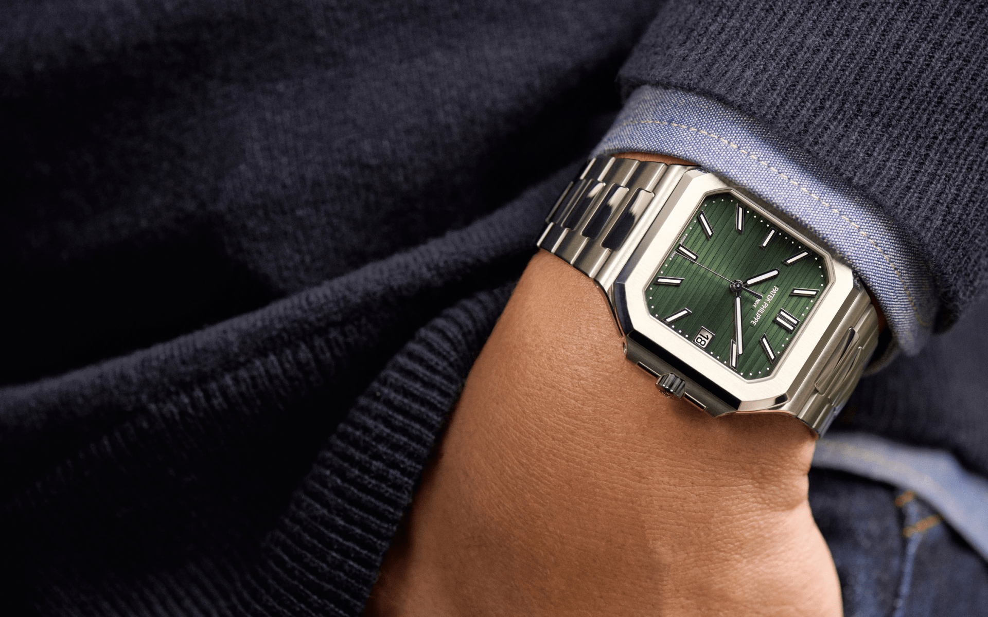 A close up of the green-faced Patek Philippe 5821/1A Cubitus watch being worn on a male wearing a light blue sweater and a dark blue sweater. The hand with the watch is being tucked into a jean pocket. 