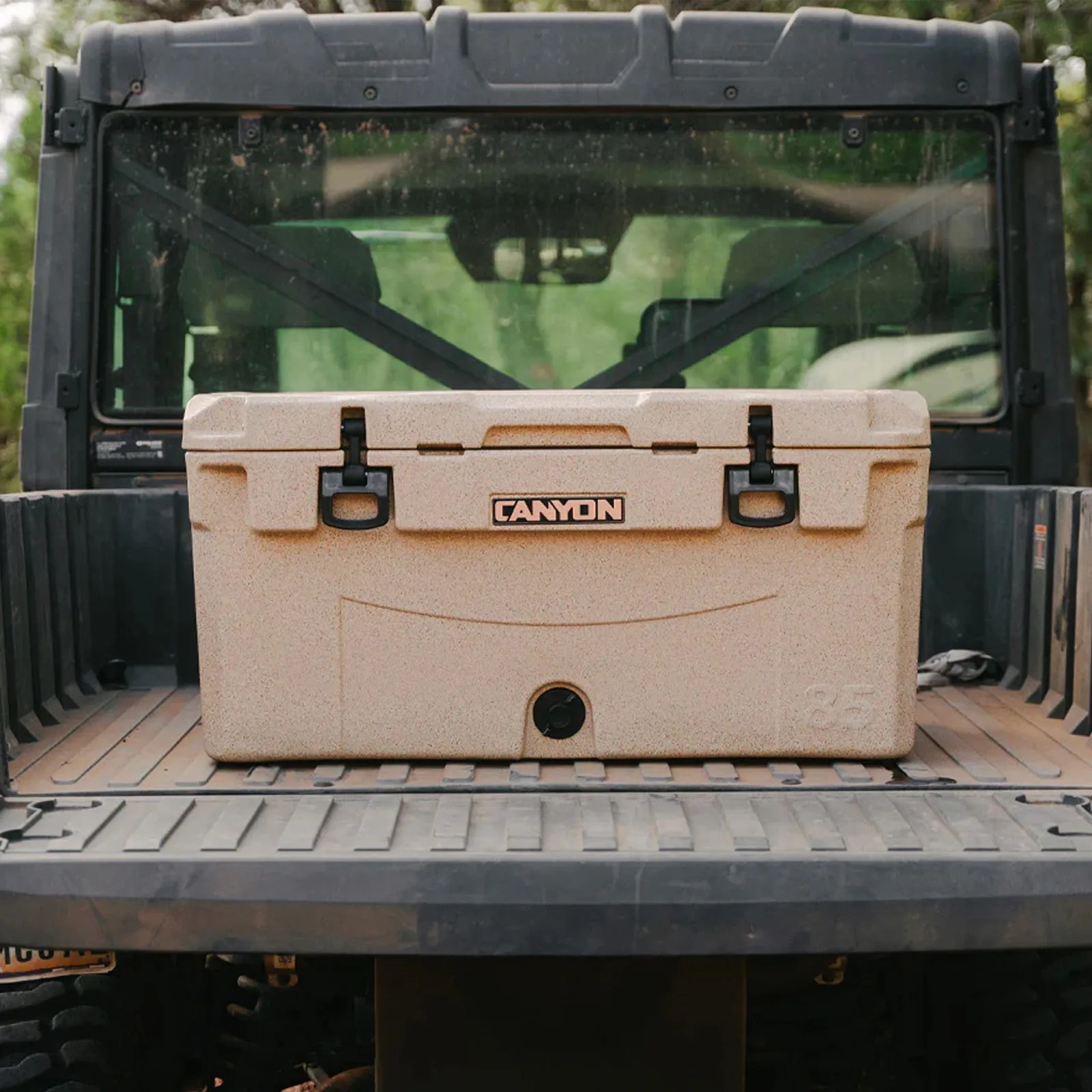 An image of a Canyon Cooler is light tan sitting in the back of a dirty ATV pickup bed. The cooler is shown from head on and fully closed.