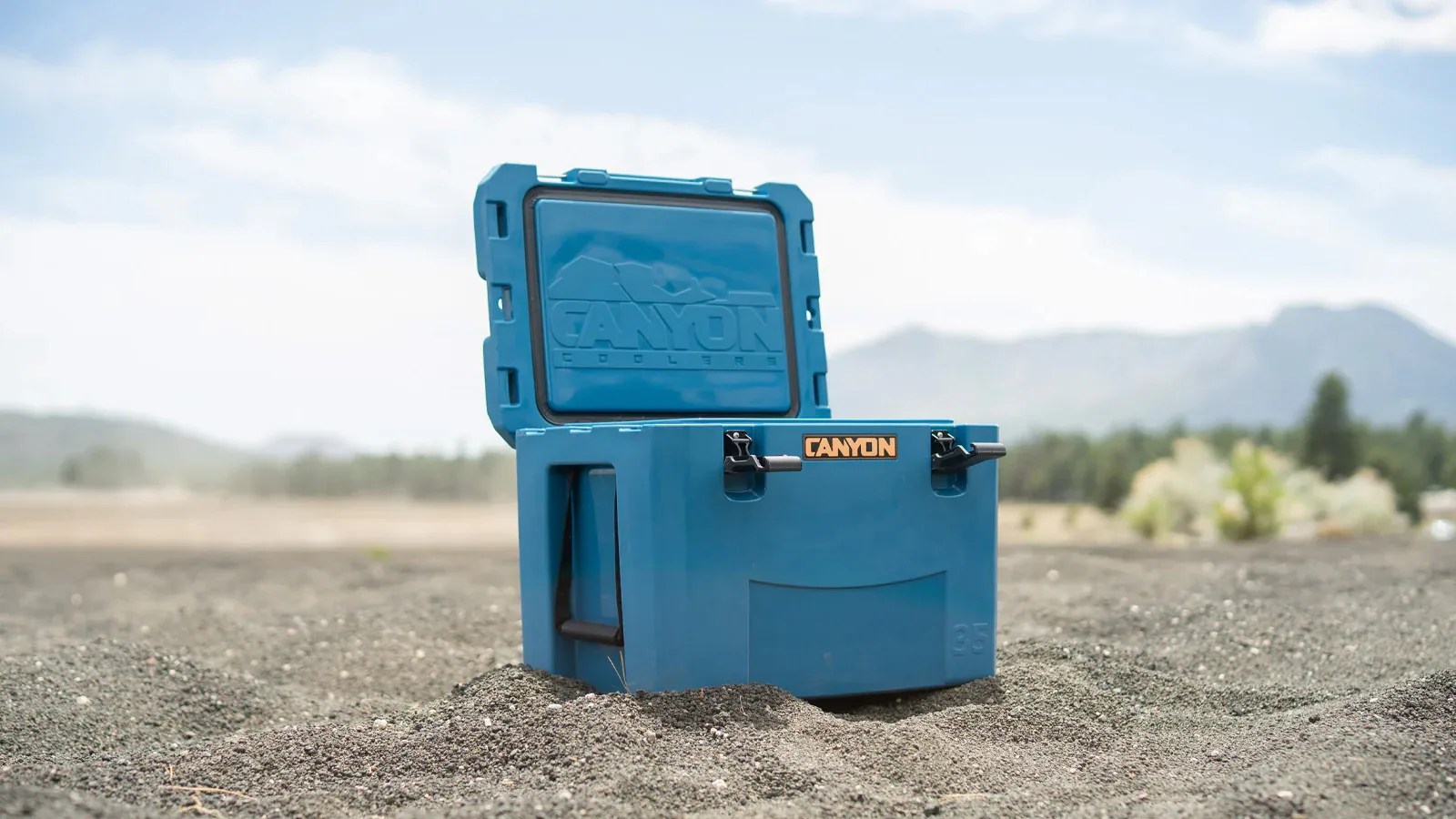 A Canyon Cooler in blue shown open and angled to the side sitting on a dark sandy bit of ground. Mountains and trees are shown blurred in the far background.