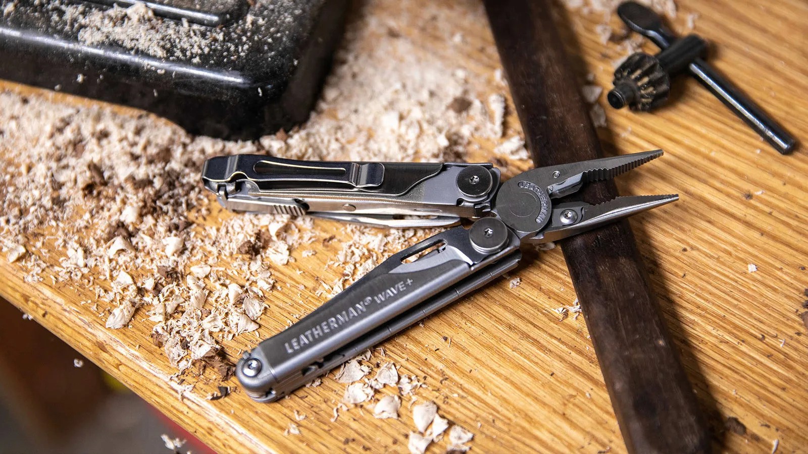 A Leatherman Wave+ multi-tool shown open in plier mode sitting on a wooden work bench surrounded by wood shavings. It's also sitting on top of a metal ruler