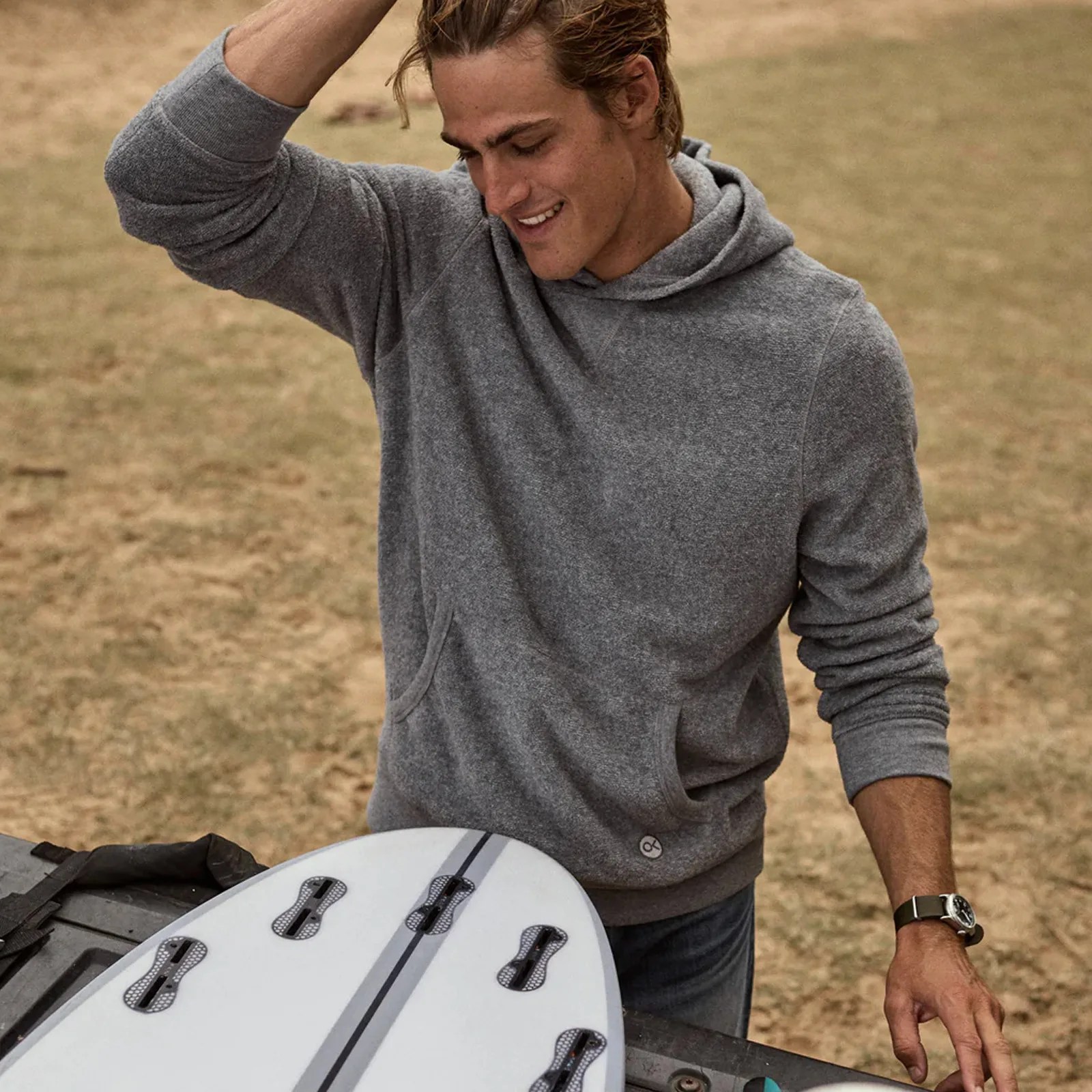 A man wearing Outerknown's Hightide Pullover hoodie in grey. He is running his hand through his hair looking at a surfboard in a pickup truck