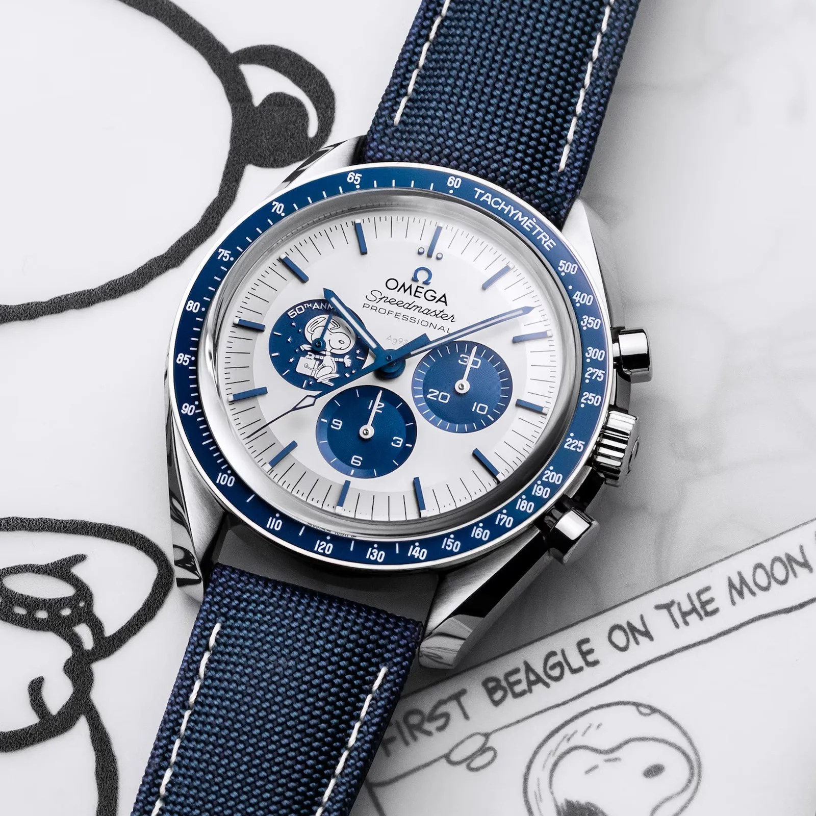 The Omega Speedmaster Annniversary Series "Silver Snoopy Award" watch shown at an angle laying on a newspaper cartoon featuring Snoopy. The Watch is white with blue marking on the dials and subdials and outer dial. It also features a blue strap. 