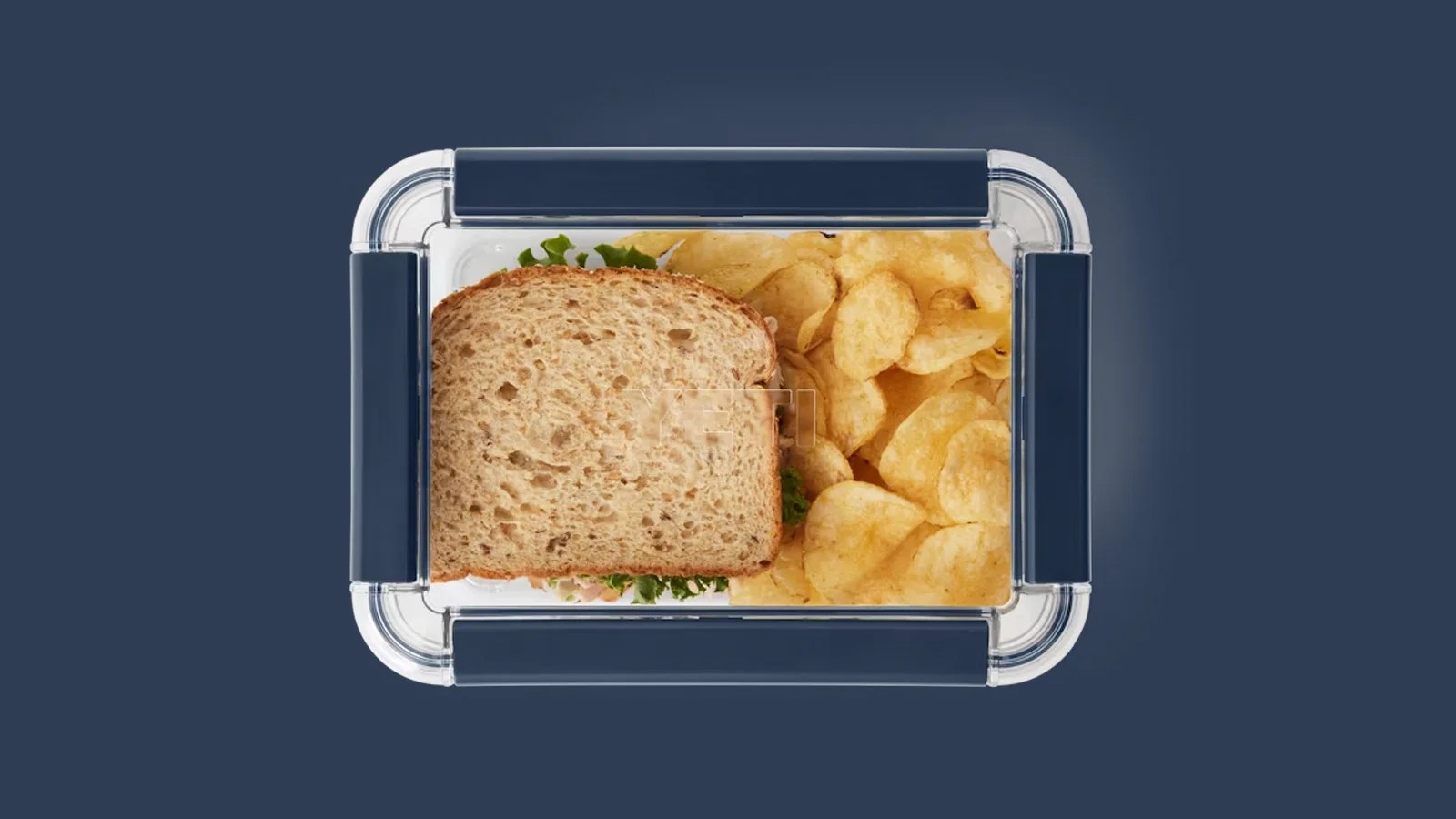 An image of Yeti's food storage container shown from above and closed. The container is holding a sandwich and potato chips. The container is shown against a solid navy blue background that matches the navy colored closure snaps on the food storage container.