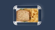 An image of Yeti's food storage container shown from above and closed. The container is holding a sandwich and potato chips. The container is shown against a solid navy blue background that matches the navy colored closure snaps on the food storage container.