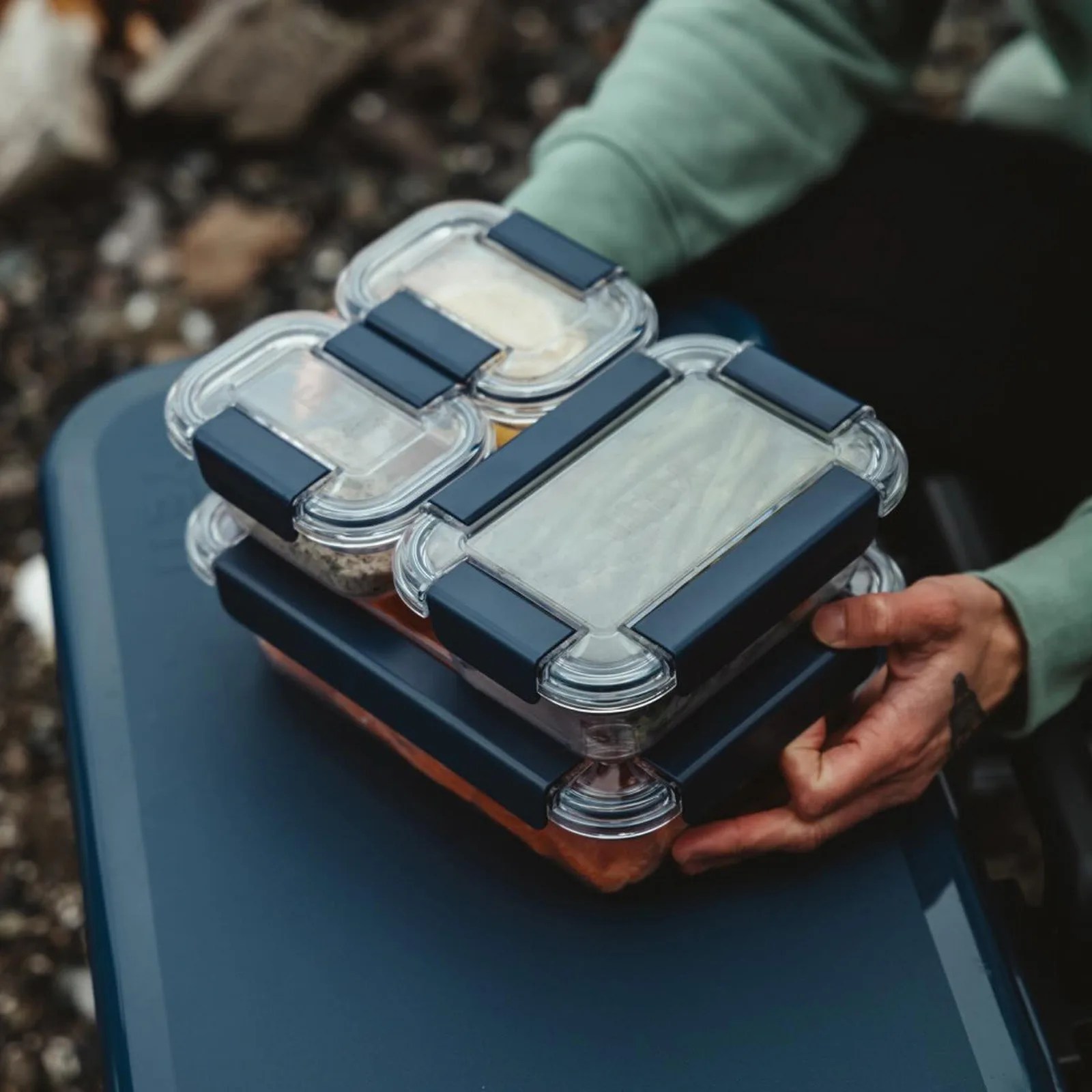 A large, medium and two small Yeti food storage containers shown stacked and nested together being held by a pair of hands. The food storage set is resting on top of a navy blue Yeti cooler that appears to be resting on a blurred gravely campsite.
