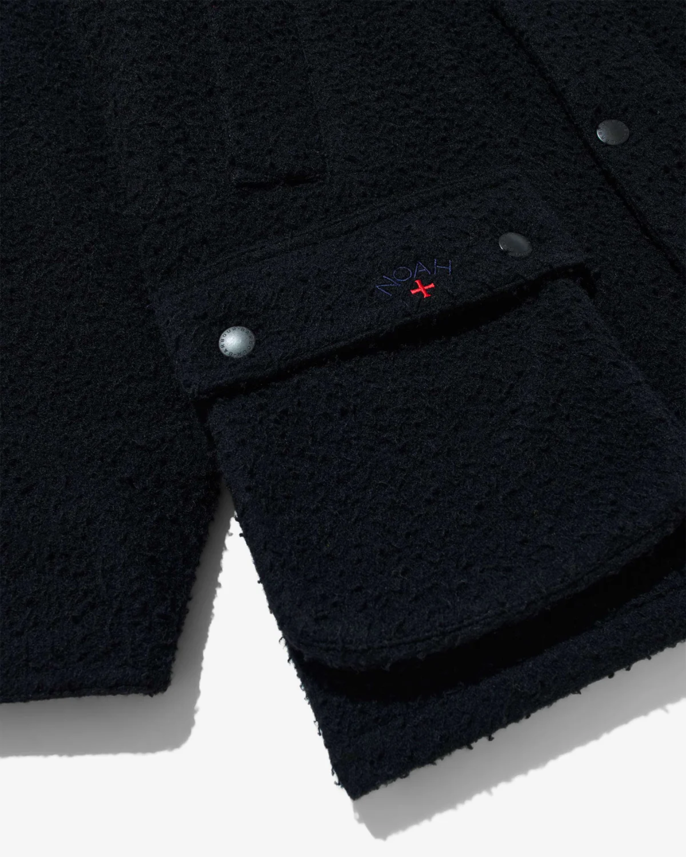 A close up image of the lower right corner and pocket area on the Barbour x Noah Wool Bedale Jacket in black. The jacket is lying flat on a light grey background. The image highlights the inclusion of Noah’s logo and red cross on the left hand pocket flap of the jacket
