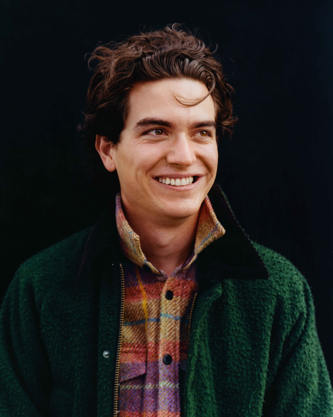 The Barbour x Noah Wool Bedale jacket being worn on a smiling male model shown from the mid torso up. The jacket is in the dark green color and unzipped. The model is wearing a bright purple and yellow plaid shirt underneath and smiling while looking off into the distance towards the right. 