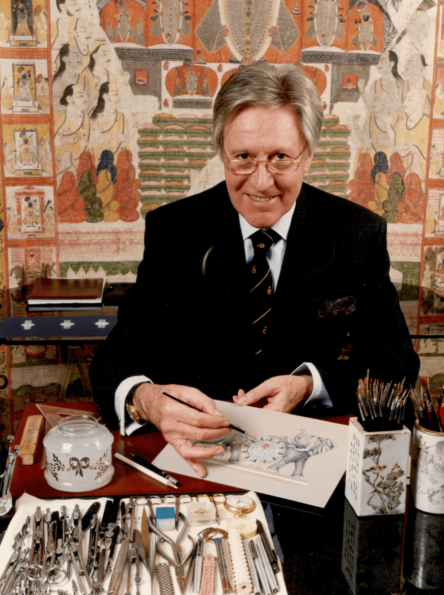 An image of Gerald Genta as a younger man sitting at a desk covered in watch tools. Genta is smiling for the photo. Is hand is holding a paint brush which he is using to paint what looks like a watch dial. Genta is wearing a blue blazer and a dark navy tie with red diagonal stripes and symbols. Behind Genta is a wall covered in busy wallpaper. 