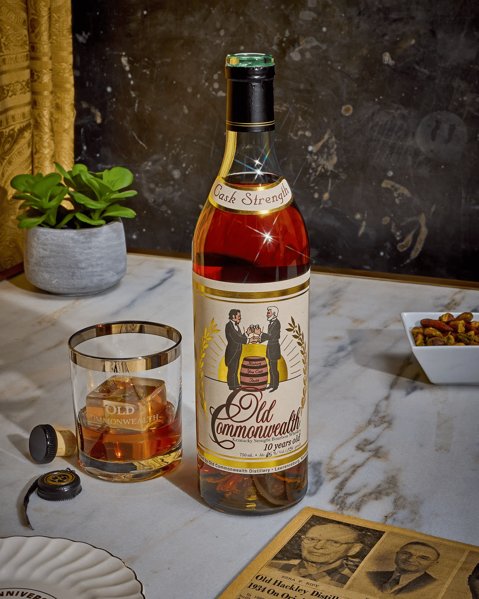 A bottle of Old Commonwealth Bourbon sitting to the right of a glass of whiskey partially filled wiht whiskey. The bottle and glass are sitting on a marble counter top The table also holds a saucer, newspaper listing, a plant and a dish of nuts