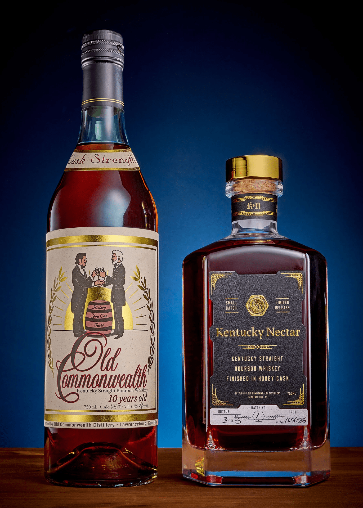 The revived version of Old Commonwealth bourbon shown next to a bottle of Kentucky Nectar also made by the same distillery. Both bottles are sitting on a wooden table in front of a blue background that fades to black at the edges. 