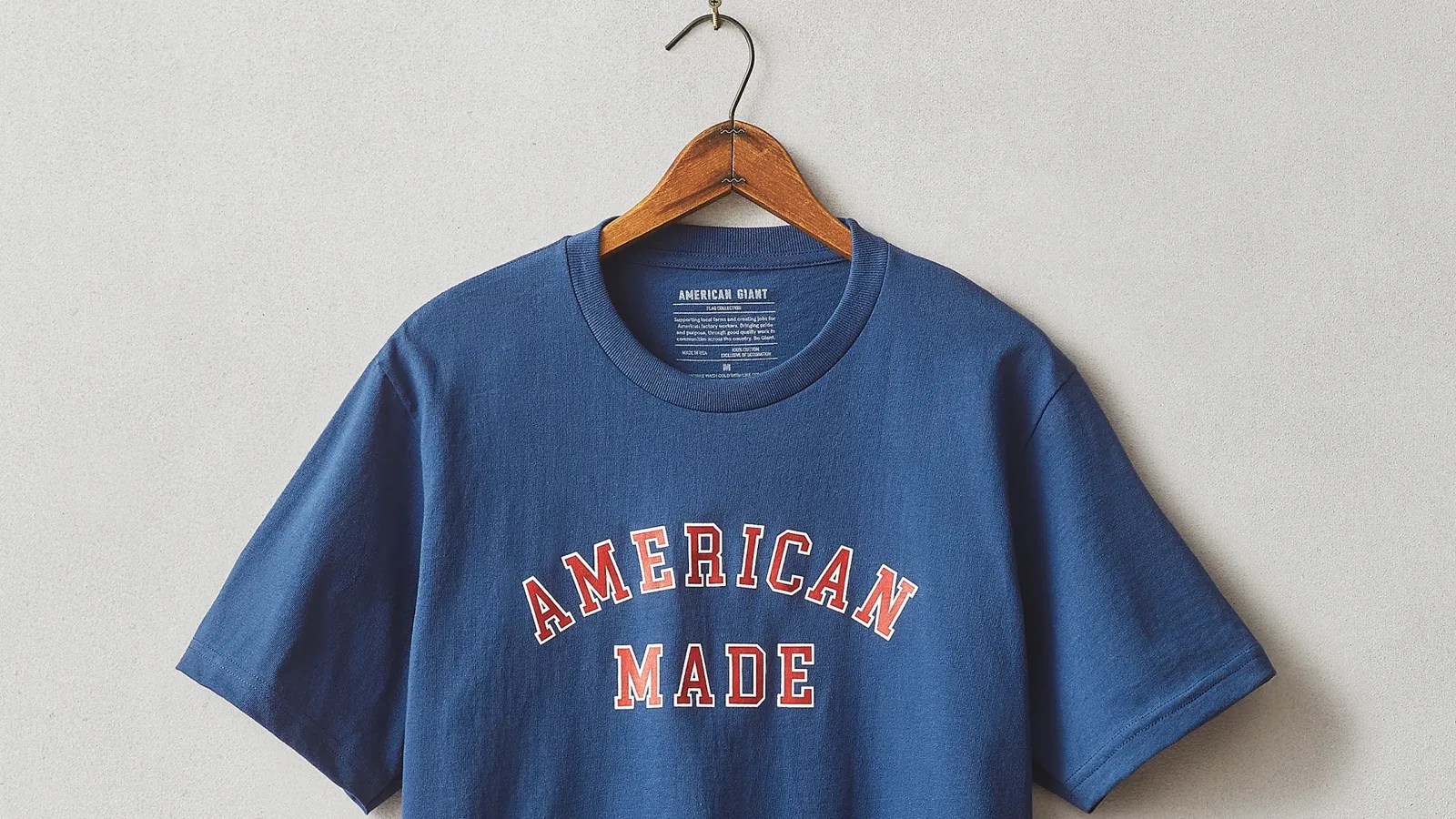 An American Made T-Shirt from Walmart and American Giant hanging on a coat hanger that is hanging off a nail. The T-Shirt is blue with the words "American Made" in red written across the chest. The shirt is shown hanging with the sleeve, neck and chest area visible. The background is a light grey wall. 