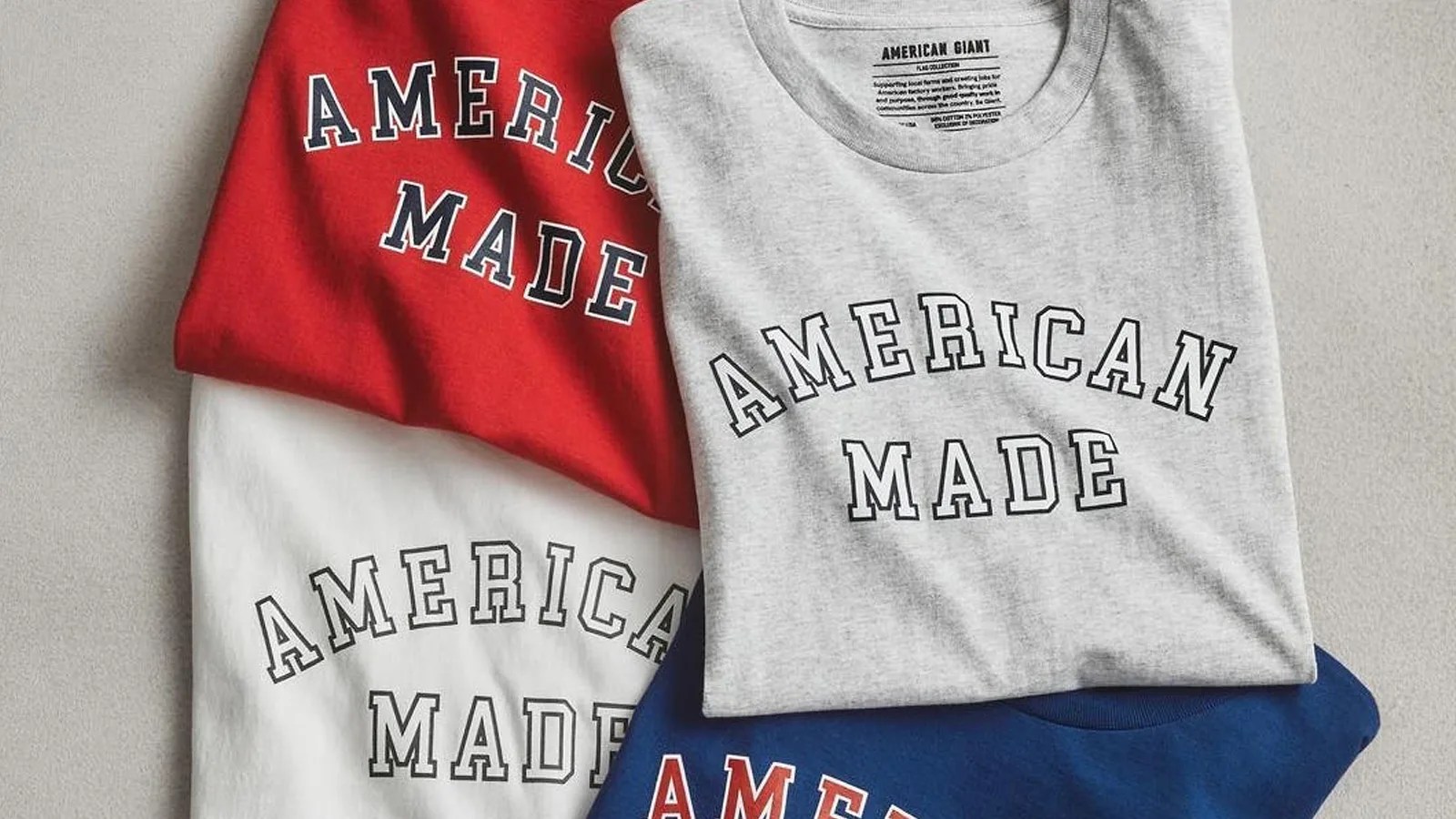 Four t-shirts with the words American Made shown on the chest stacked in a grid with some of their edges overlapping. One t-shirt is red, one is blue and two are grey. They are sitting on a light grey surface.