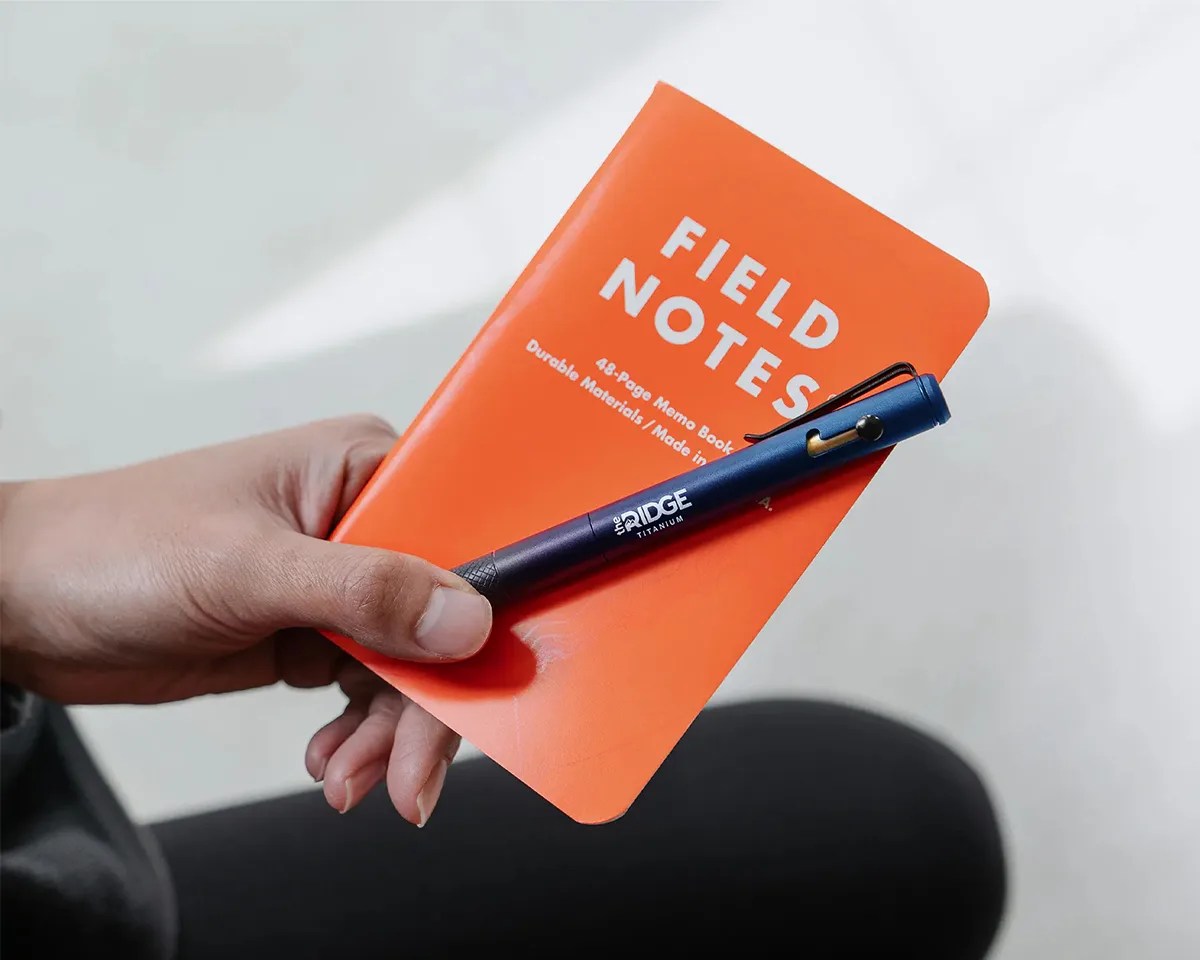 The Ridge Bolt-action pen held in a person's hand next to a Field Notes notebook