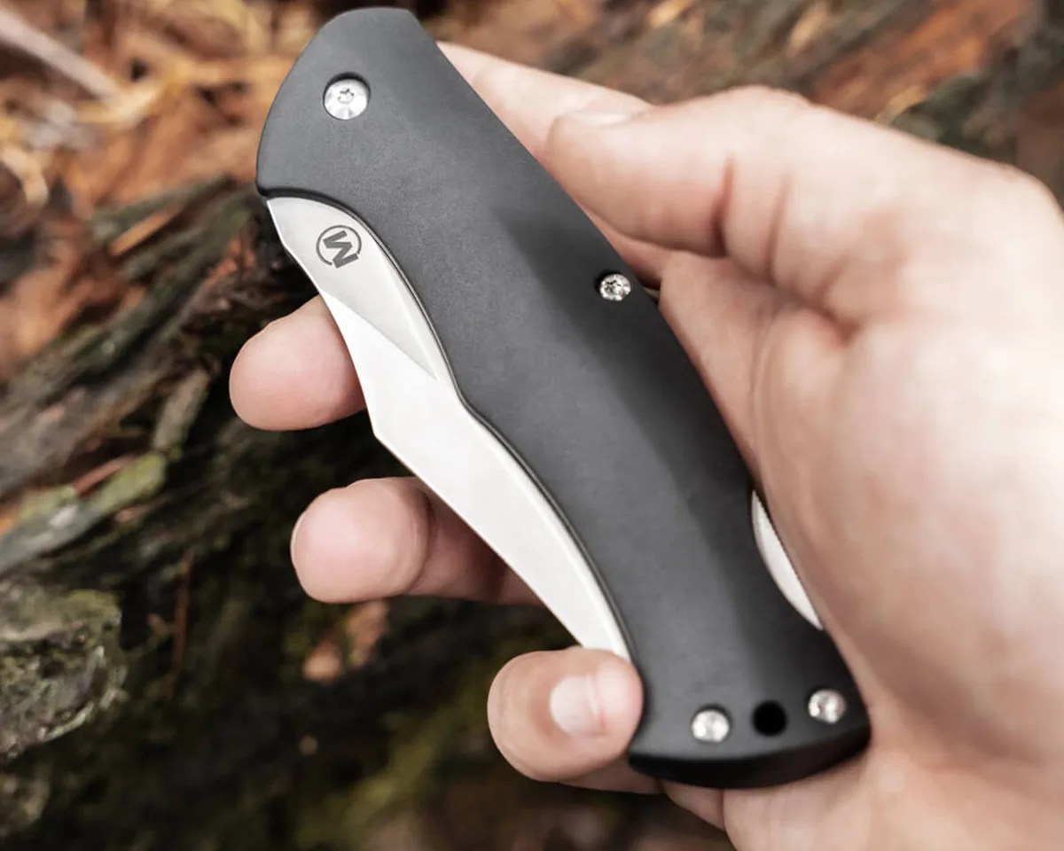 A hand holding closed Boker Magnum Echidna knife over forest floor background