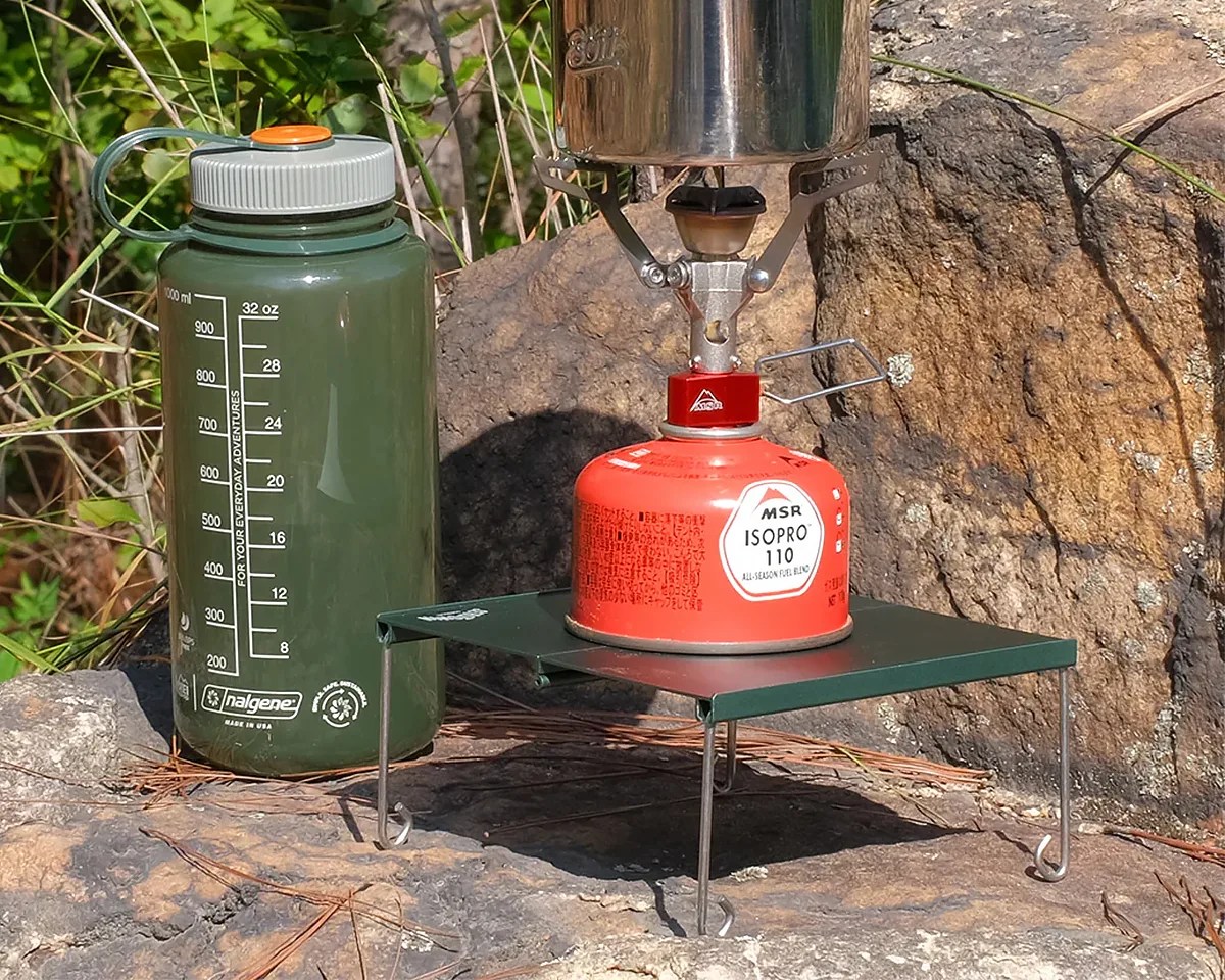 Serotonin Outdoors Pocket Mini Table with MSR camp stove on top of it next to Nalgene bottle on a rock in the woods