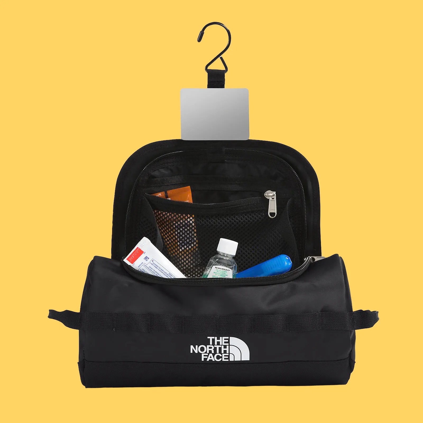 The North Face Base Camp Travel Canister shown open to reveal the integrated hanger and mirror. It's also shown filled with small toiletries. The black bag is setting against a bright yellow background.