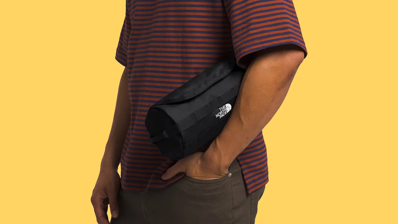 The North Face Base Camp Travel Canister bag being held in the crook of the fore arm of a male model facing towards the left of the image. The model is wearing a red and blue striped shirt. The bag is black. The background of the image is a bright yellow.