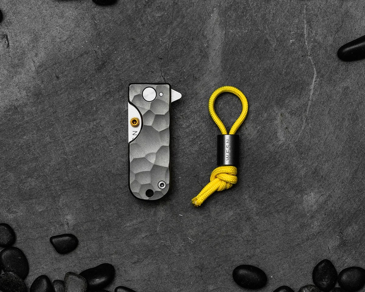 Closed WESN Ridgeback Microblade on slate with yellow lanyard surrounded by black pebbles