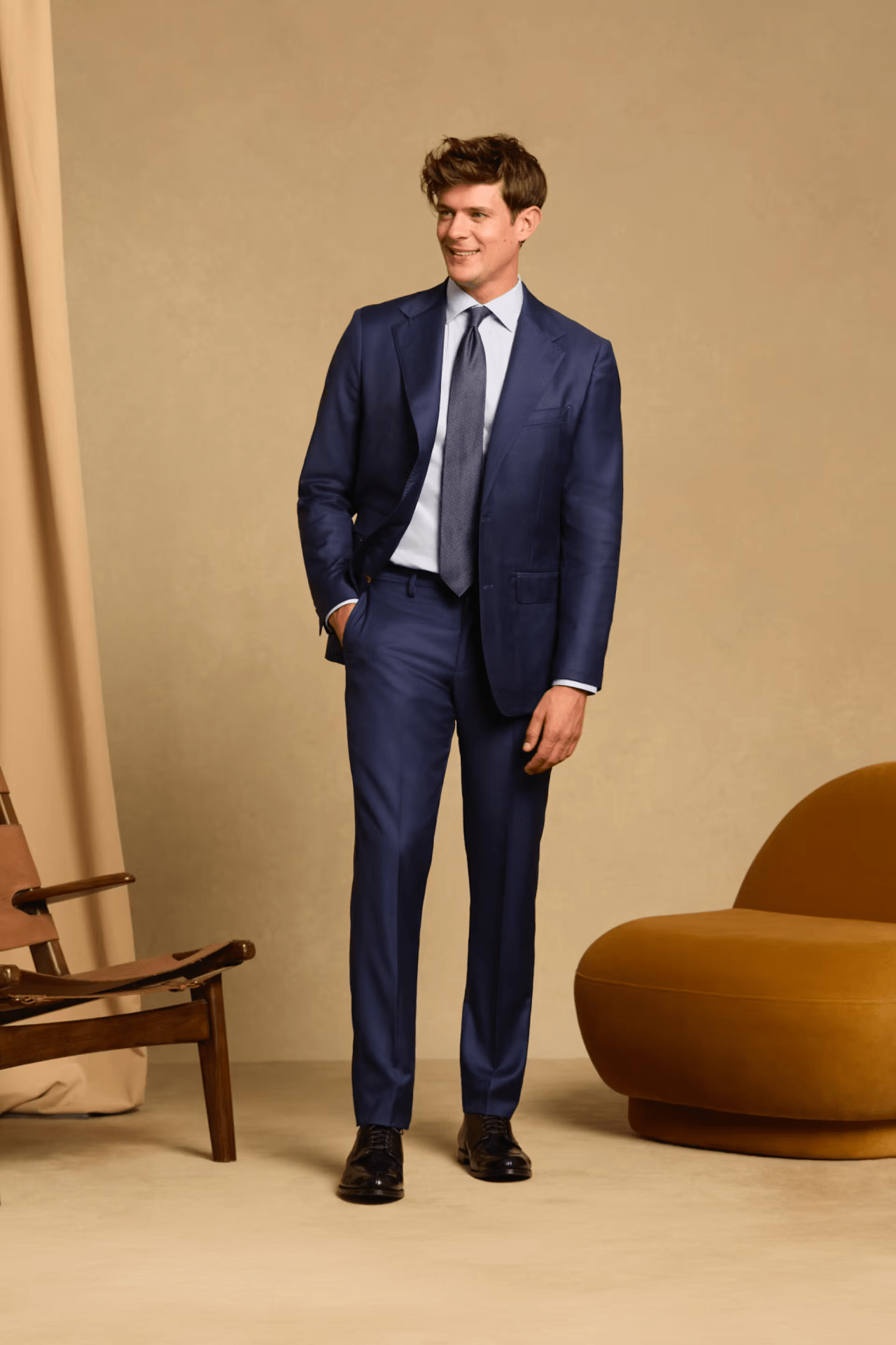 A white male wearing the Bonobos Empire 150 suit in navy with his right hand in his suit pocket while looking to the side. He is standing in a tan colored room between two stylish lounge chairs. One is made of wood. The other is puffy and made from an orange fabric.