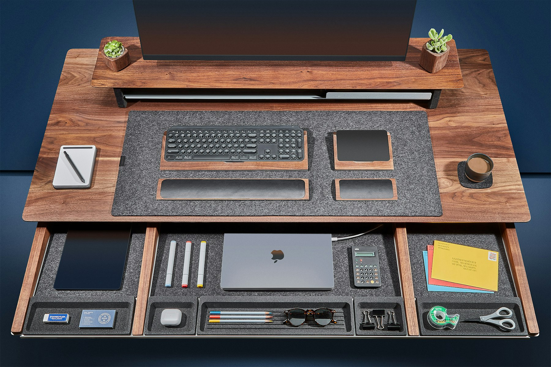 The a dark walnut version of the Grovemade Desk shown from above with the main drawer open to reveal various compartments containing a variety of office accessories including a laptop, pens, paperclips, scissors, tape and tablet.
