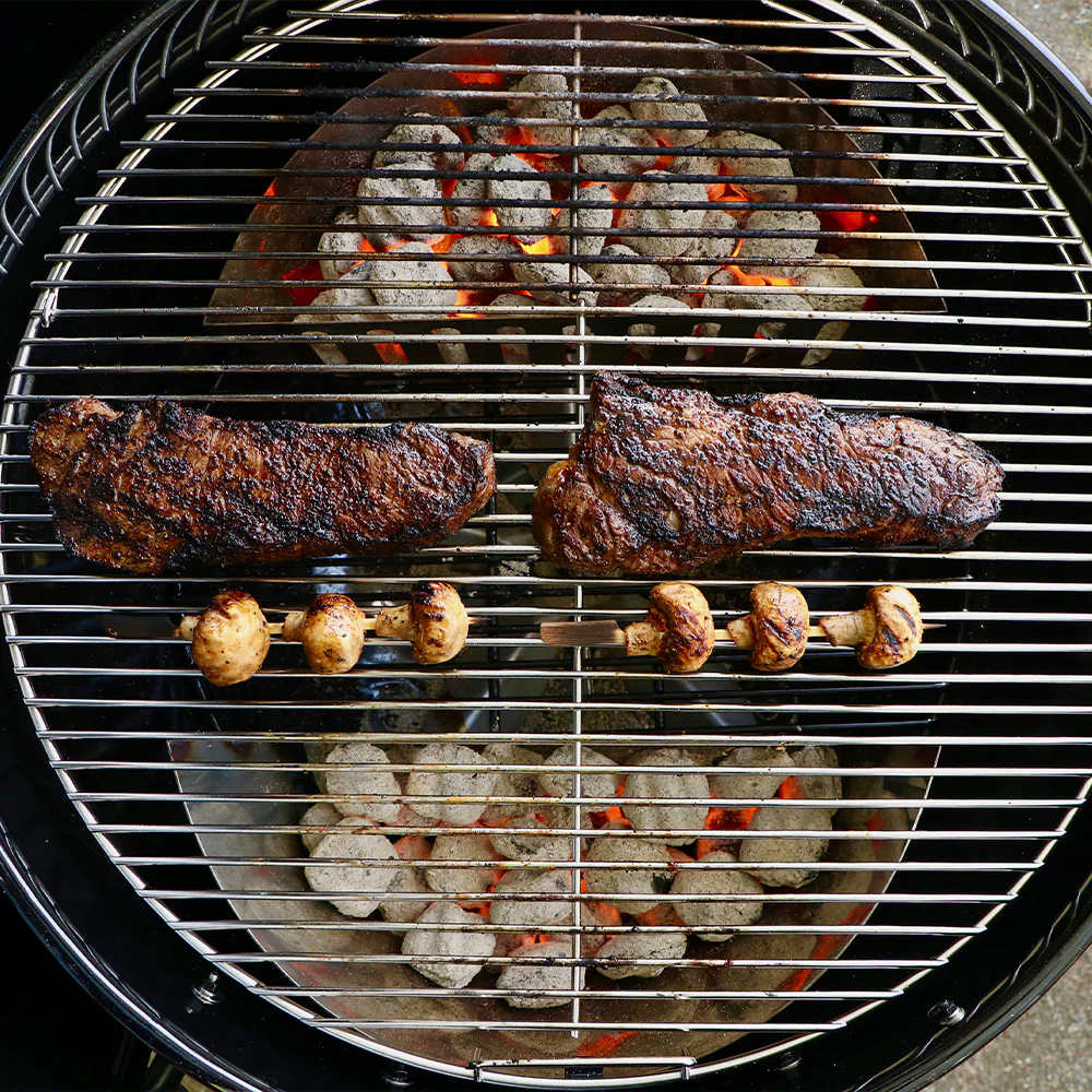 A top down photo of an active charcoal grill featuring SnS Grill baskets sitting underneath a charcoal grill grate. Steaks and mushrooms are grilling on top of the grates.