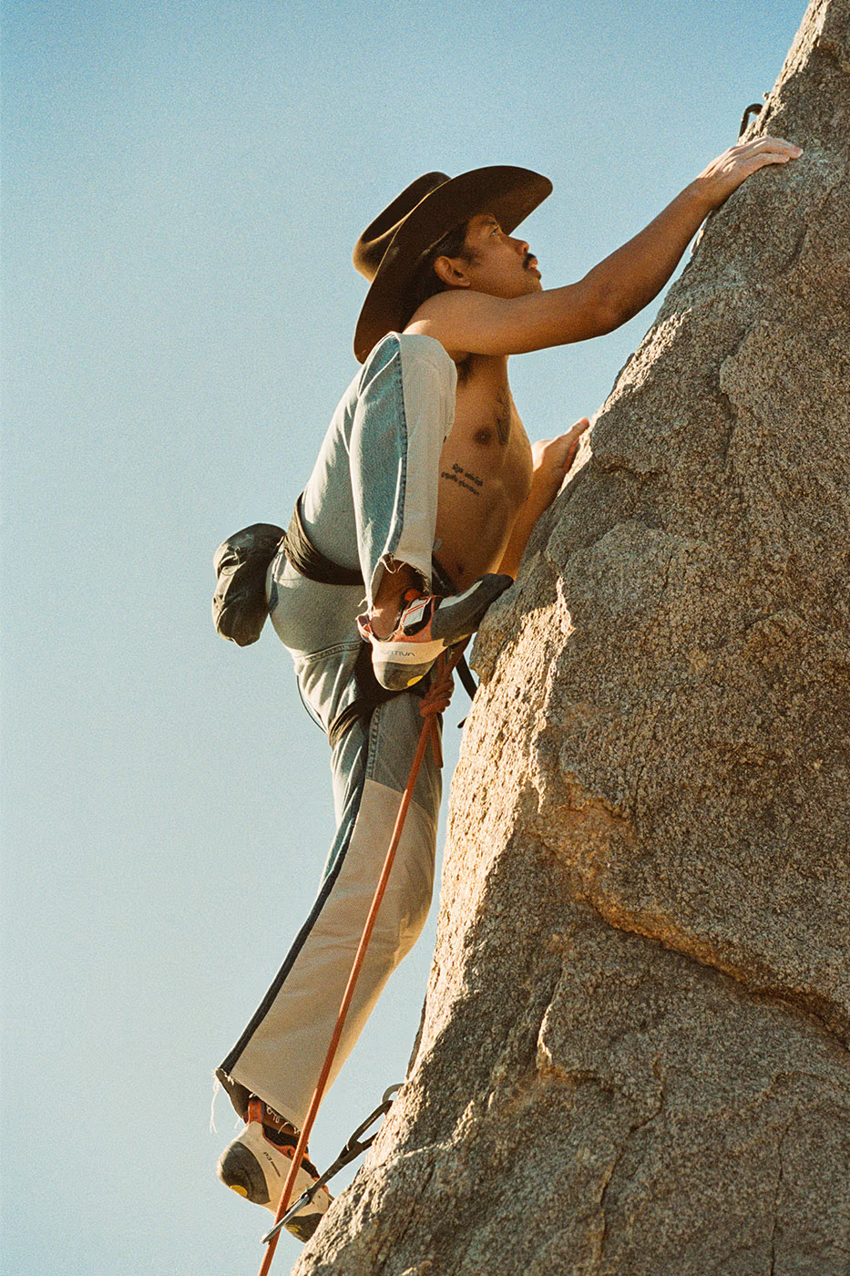 A shirtless man climbing a rock face wearing a cowboy hat and the Levi's 501 Re-Possessed Climb Jeans along with climbing shoes. 