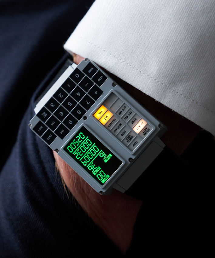 The Apollo Instruments DSKY Moonwatch being worn on a man's wrist. The man's hand is sitting inside the pocket of a pair of navy blue pants. The watch's display is lit up to show the various glowing yellow buttons and bright green screen. 