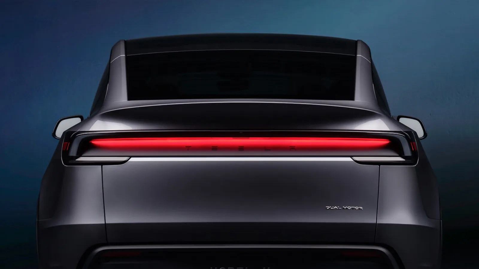 A close up of the rear of the updated Tesla Model 7 for 2024. The car is a grey color and the rear taillight bar is shown lit. The car is sitting against a dark blue background.