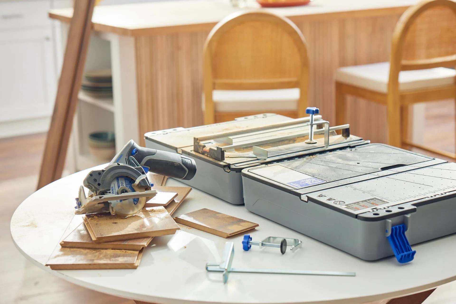 Dremel Blueprint portable table saw shown sitting on a table alongside some cut pieces of wood. In the background is a blurred kitchen island with bar chairs. 