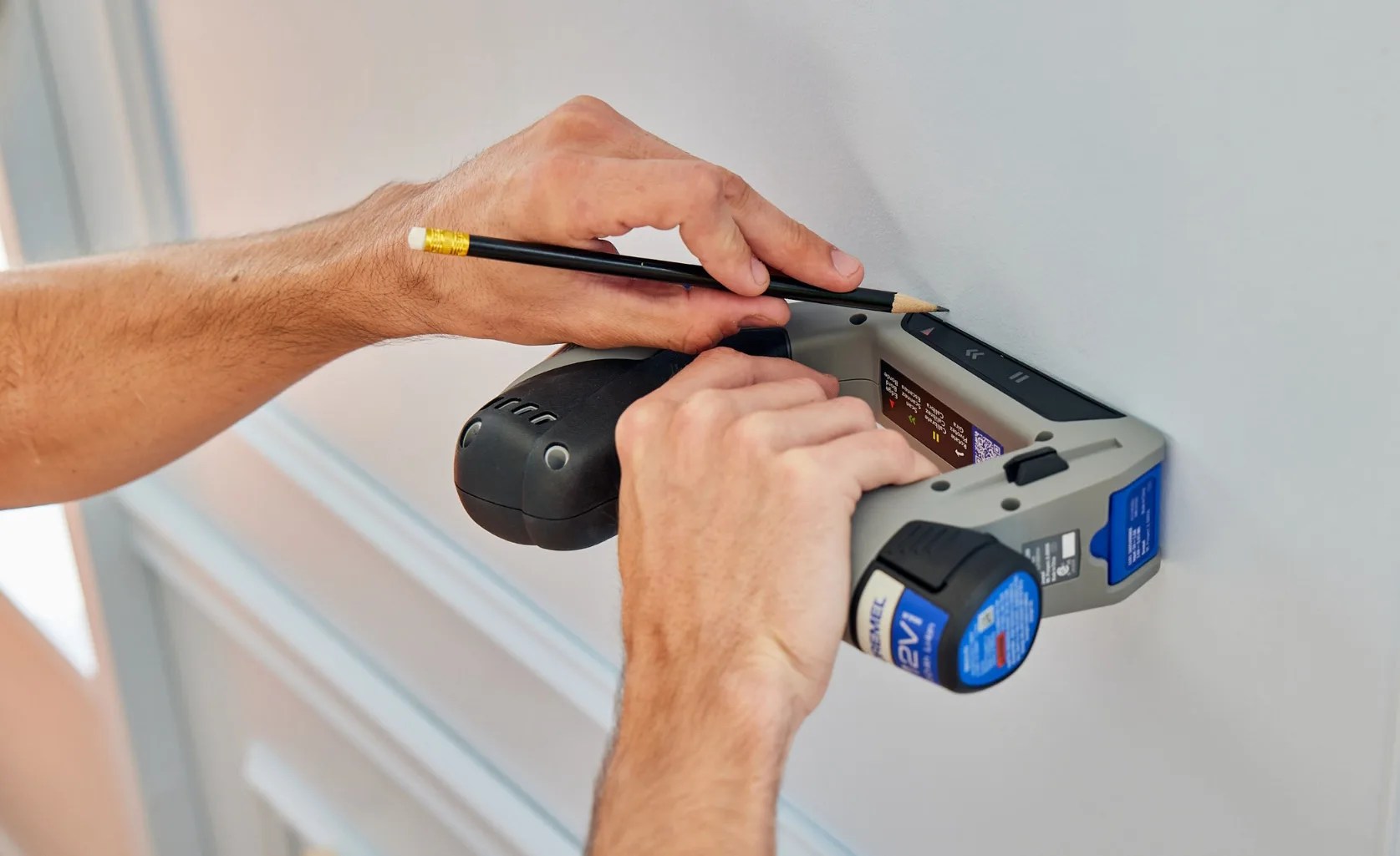 The Dremel Blueprint Multi-Drill shown being pressed flush against a white wall and turned to its side to highlight the device's integrated stud finder functionality. A pair of hands are shown along with the drill. One hand is holding the drill against the wall. The other hand is marking a spot on the wall with a black pencil.