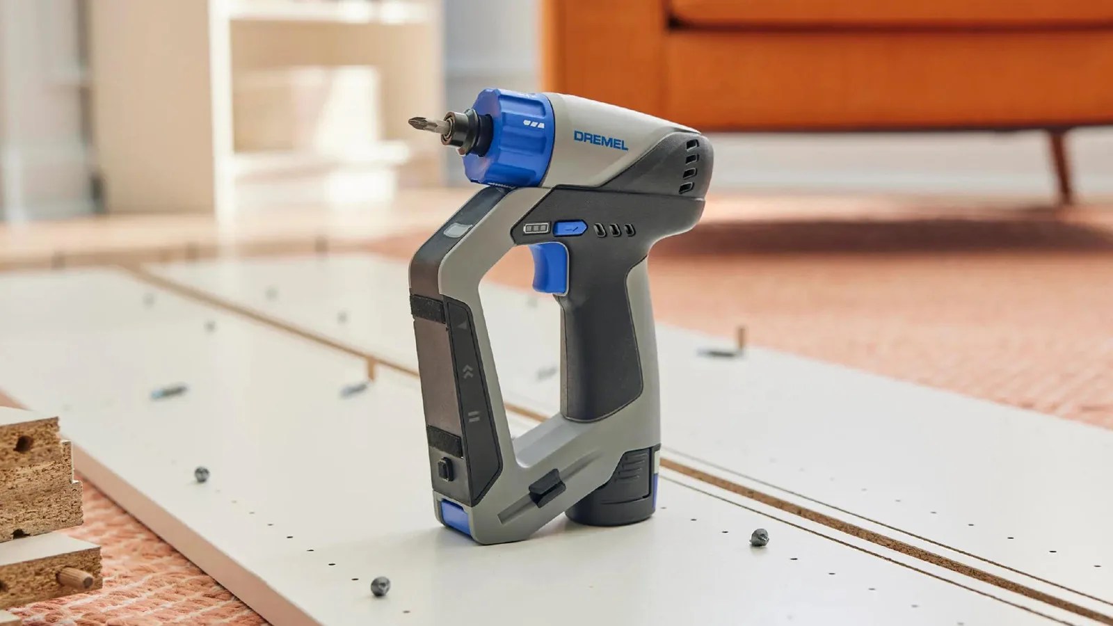 The Dremel Blueprint Multi-Drill shown sitting on a room floor sitting on top of two white planks of wood that appear to be furniture parts being assembled. The bottom feat of a credenza appears in the background