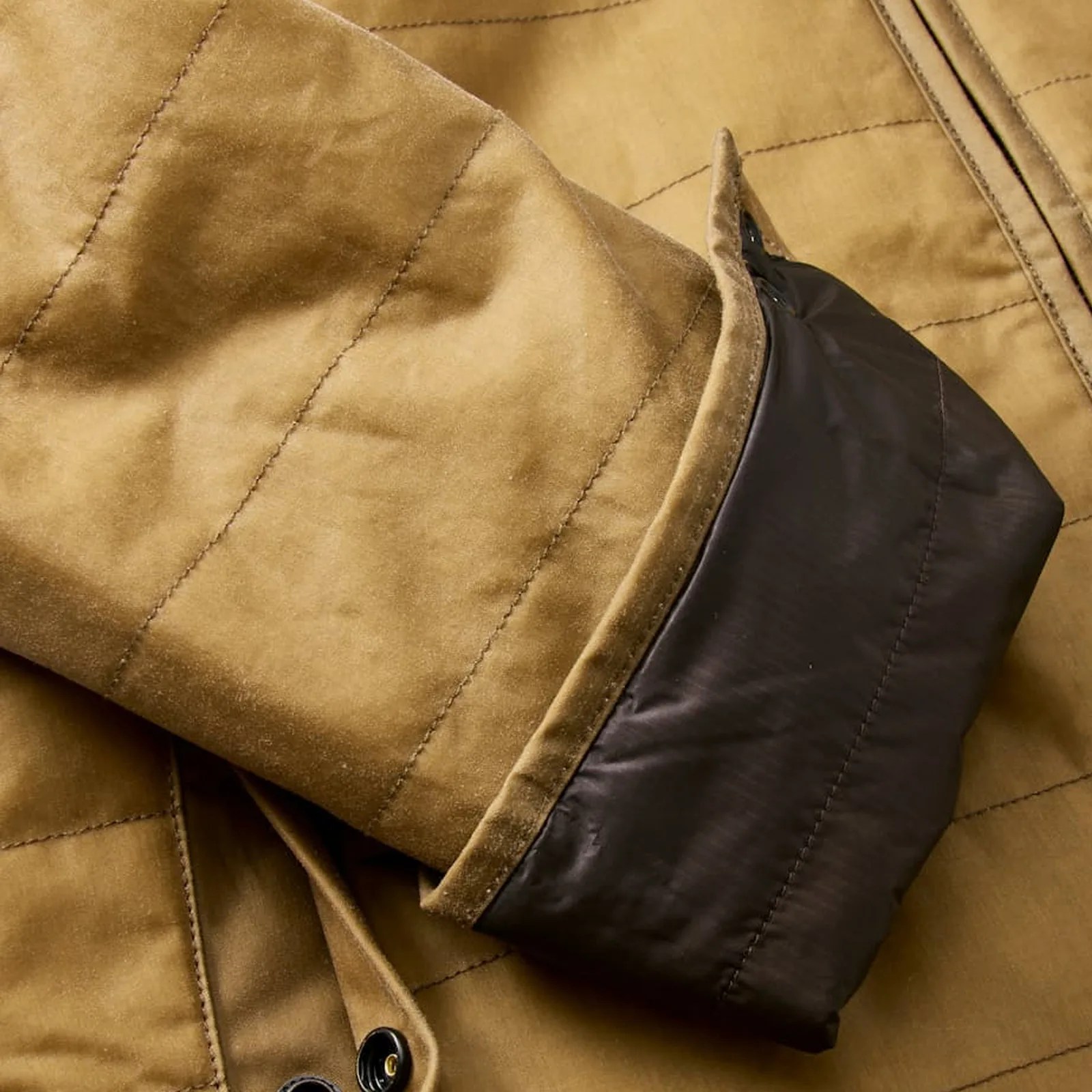 A close up of the cuff of the Flint and Tinder York Waxed Canvas Puffer Jacket. The jacket shown is a golden tan color and the close up shows the left cuff rolled up to reveal a dark chocolate brown lining.